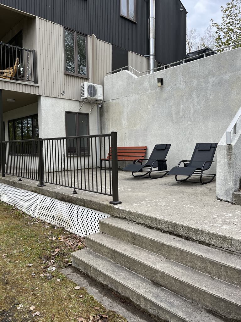 Une terrasse en béton avec deux chaises longues noires et un banc en bois, entourée d'une balustrade en métal noir, à l'extérieur d'un immeuble moderne de deux étages aux murs gris et équipé d'un climatiseur de fenêtre. Un escalier mène à un espace vert.