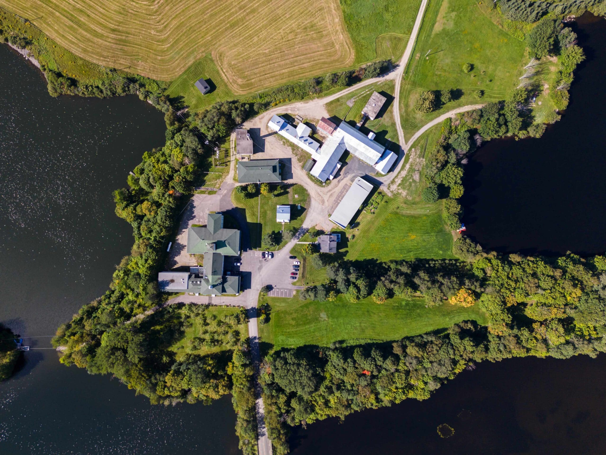 Vue aérienne d'un complexe agricole rural au bord d'un lac, montrant les bâtiments, les champs, les routes et les espaces verts environnants.