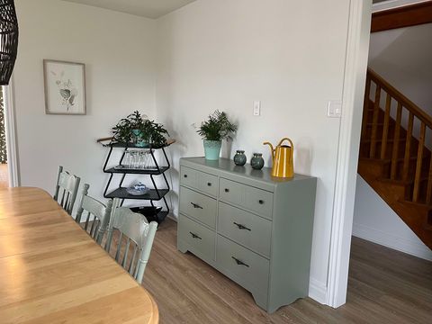 Une commode vert clair, ornée de plantes et d'objets décoratifs, se dresse contre un mur blanc près d'un escalier. Une table à manger et des chaises en bois sont au premier plan, et un support à plantes à plusieurs niveaux trône dans un coin.