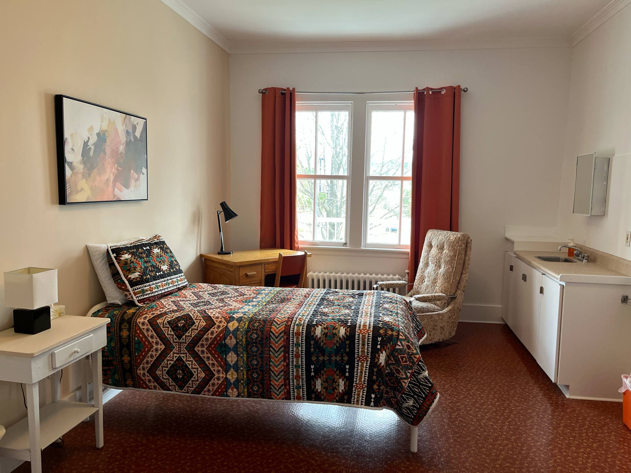 Une chambre confortable avec un lit simple, une literie à motifs colorés, un bureau en bois avec une lampe, une chaise rembourrée, deux fenêtres avec des rideaux rouges et un coin lavabo blanc avec un miroir.