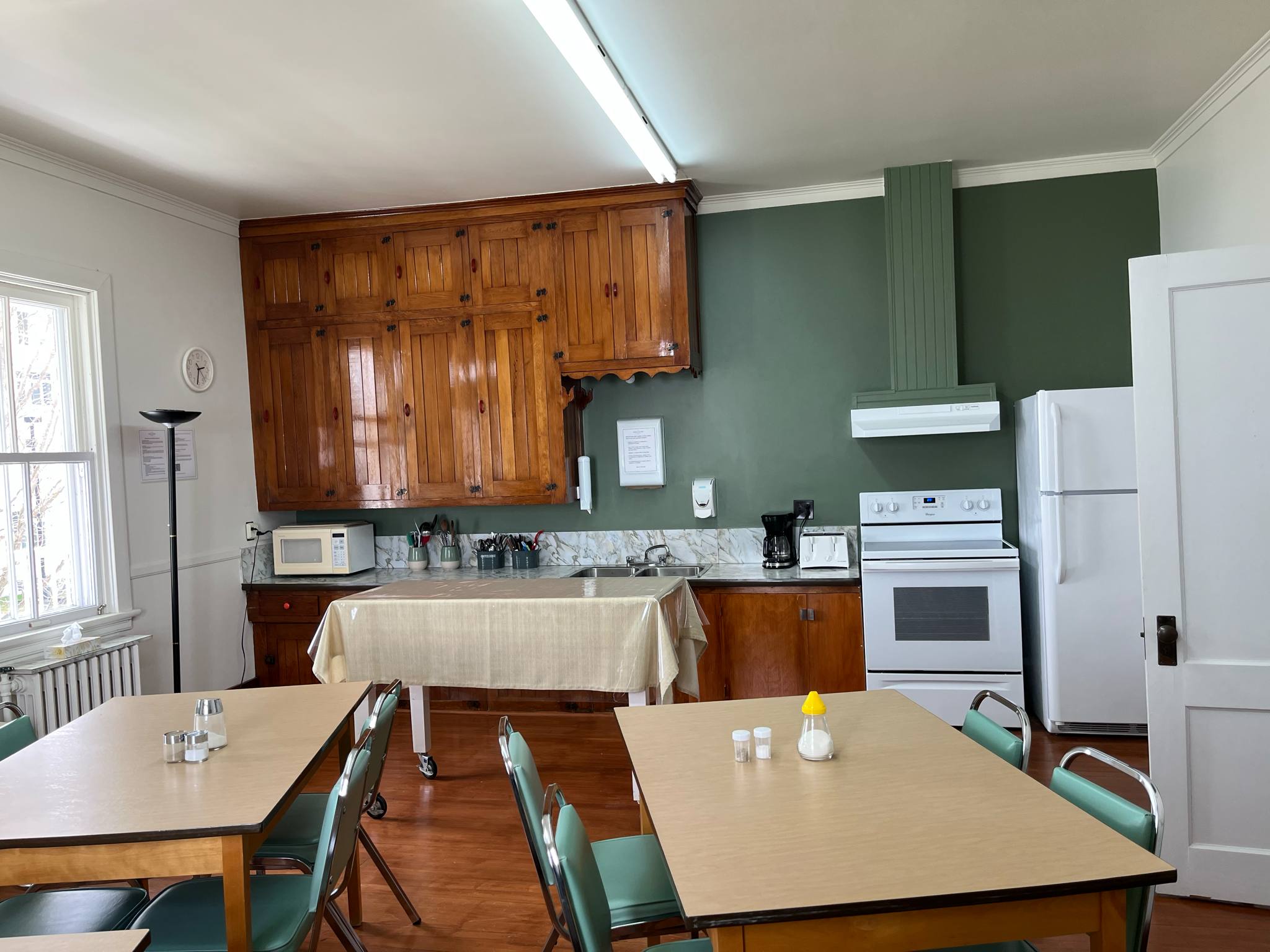 Une cuisine aux murs verts, aux placards en bois, avec une cuisinière blanche, une hotte, un réfrigérateur et un micro-ondes. Deux tables avec des chaises vertes et des condiments sont disposées au premier plan ; une table avec une nappe trône au centre.