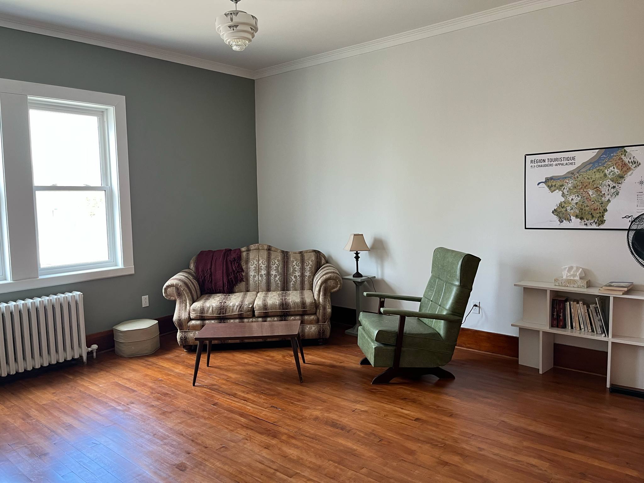 Un salon simple avec une causeuse à motifs, un fauteuil vert, une petite table en bois, un lampadaire, une bibliothèque avec des livres, une carte murale, un parquet et une fenêtre laissant entrer la lumière naturelle.