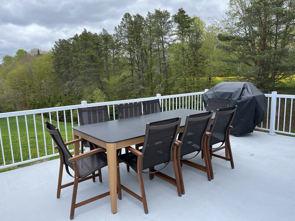 Une terrasse avec une table noire et huit chaises est installée sur une terrasse bordée d'une balustrade blanche. Un barbecue couvert se trouve à proximité. En arrière-plan, des arbres verts et un ciel nuageux.