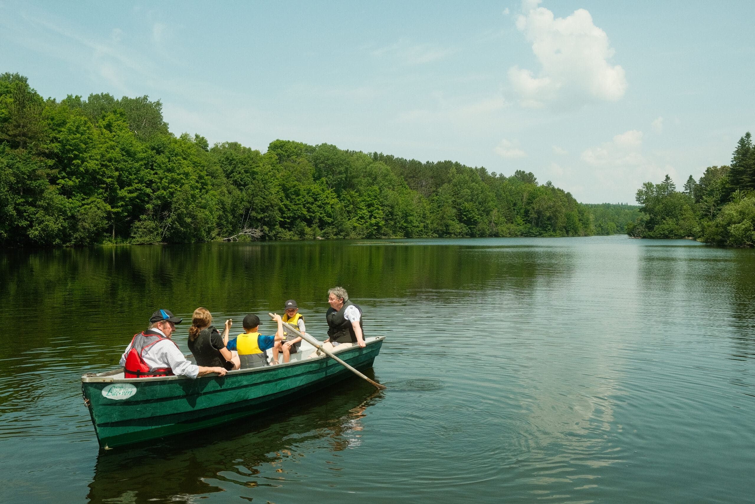 Un groupe de cinq personnes, dont des enfants et des adultes, profite d'une sereine promenade en bateau sur un lac calme entouré de forêts verdoyantes sous un ciel bleu clair.