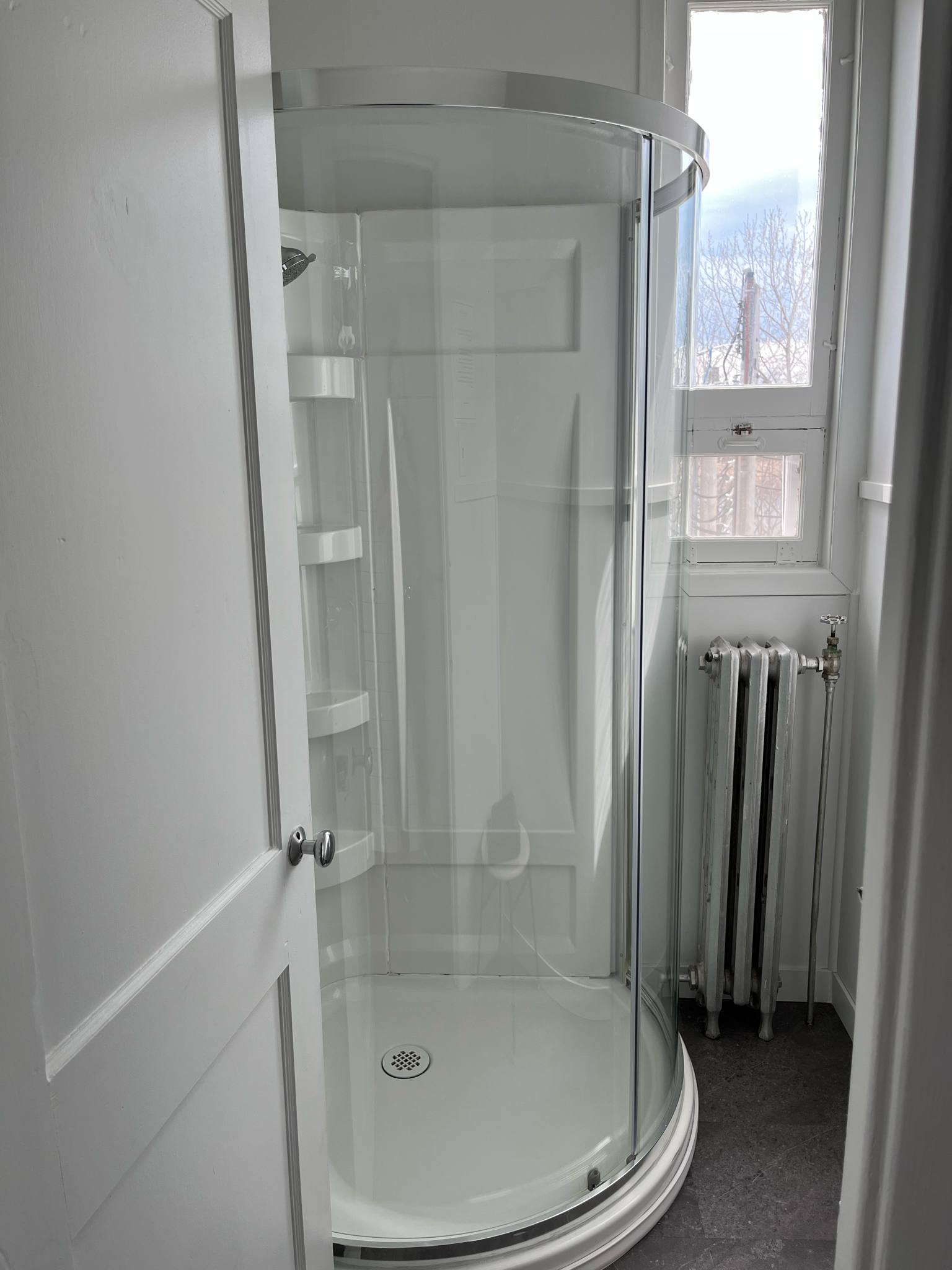 Une petite salle de bain avec une cabine de douche en verre incurvé, une porte blanche partiellement ouverte, une fenêtre laissant entrer la lumière naturelle et un radiateur sous la fenêtre. Les murs et la douche sont blancs.