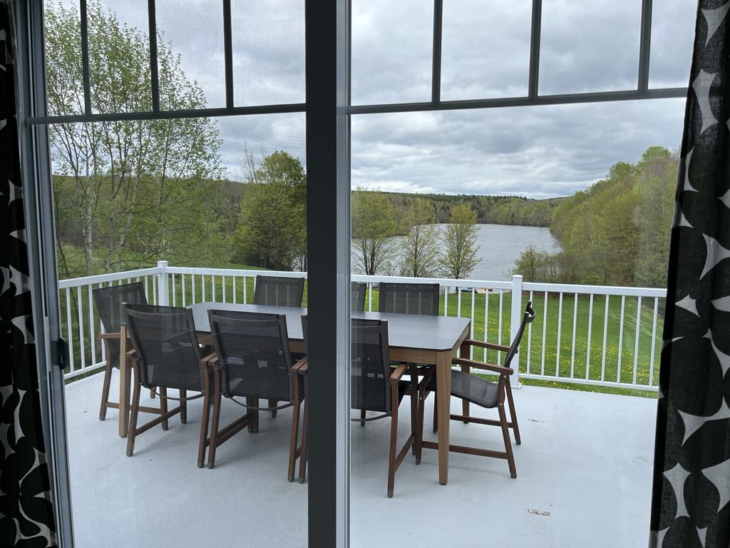 Vue à travers une porte vitrée d'un patio avec une table et huit chaises, donnant sur une pelouse verte, des arbres et un lac sous un ciel nuageux.
