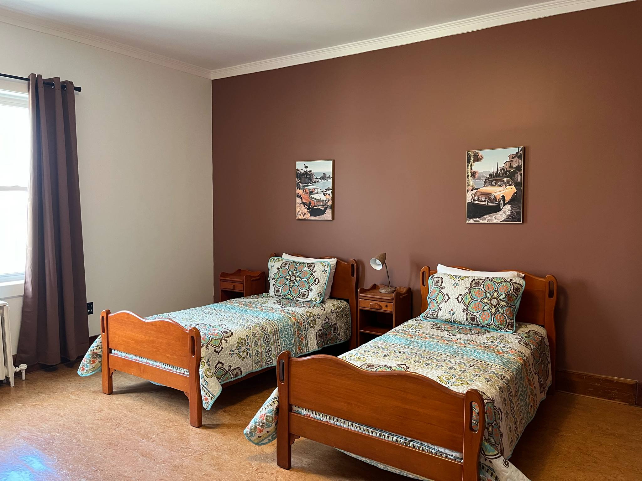 Deux lits jumeaux en bois, ornés de couvre-lits aux motifs colorés, sont disposés côte à côte contre un mur marron. Chaque lit est doté d'un oreiller et d'œuvres d'art encadrées au-dessus de la tête de lit. Une fenêtre avec des rideaux sombres se trouve à gauche.