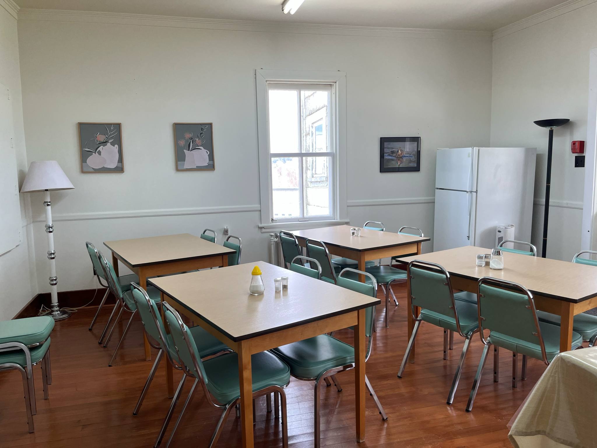 Une salle à manger simple avec quatre tables, des chaises vertes, une salière et une poivrière, deux lampes, un réfrigérateur blanc et des œuvres d'art sur des murs clairs. La lumière du soleil entre par une seule fenêtre.