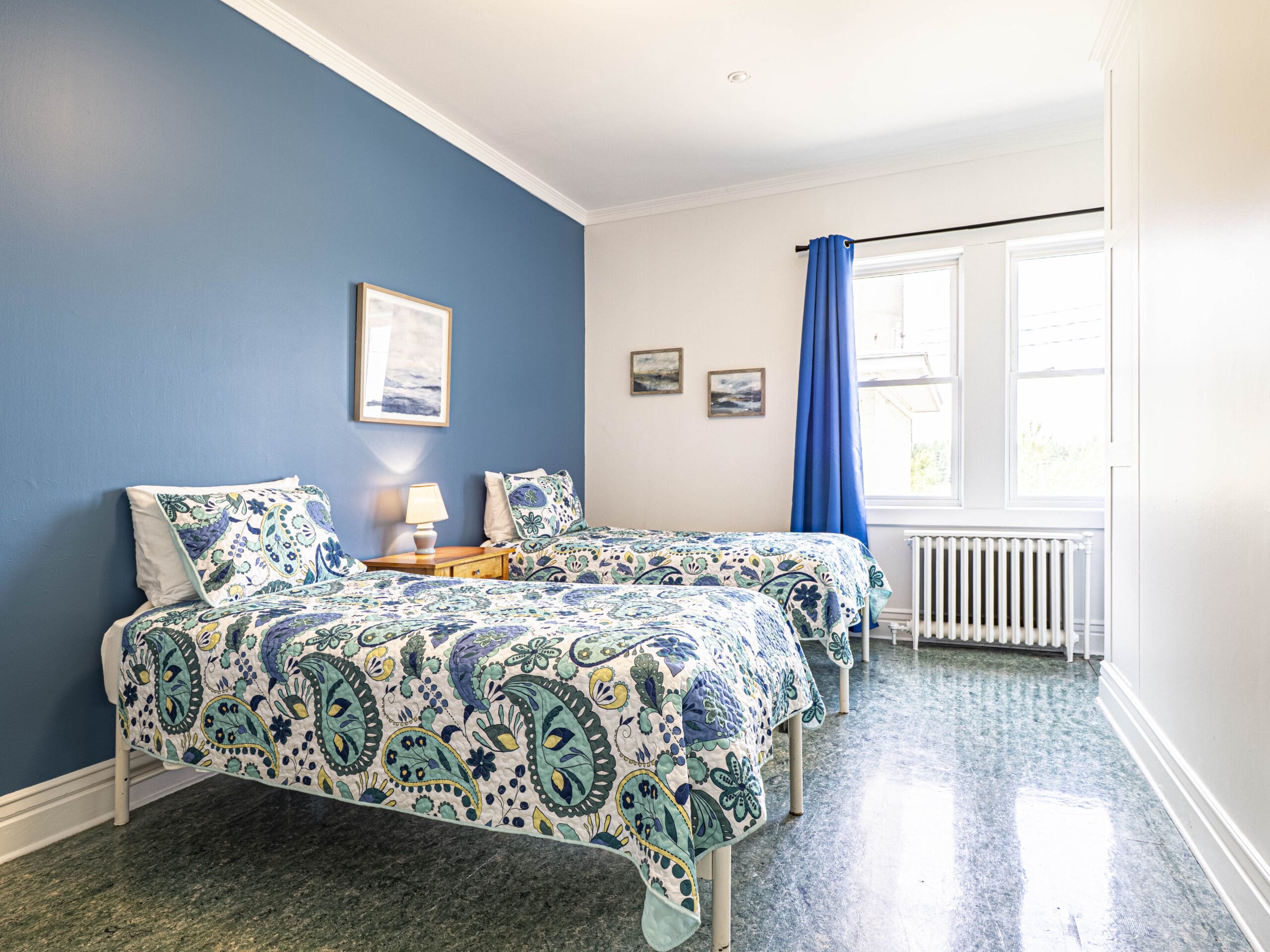 Chambre lumineuse avec deux lits simples recouverts d'une literie bleue et blanche, un mur d'accent bleu, trois tableaux encadrés, une table de nuit avec une lampe, une grande fenêtre avec des rideaux bleus et un sol à motifs verts.