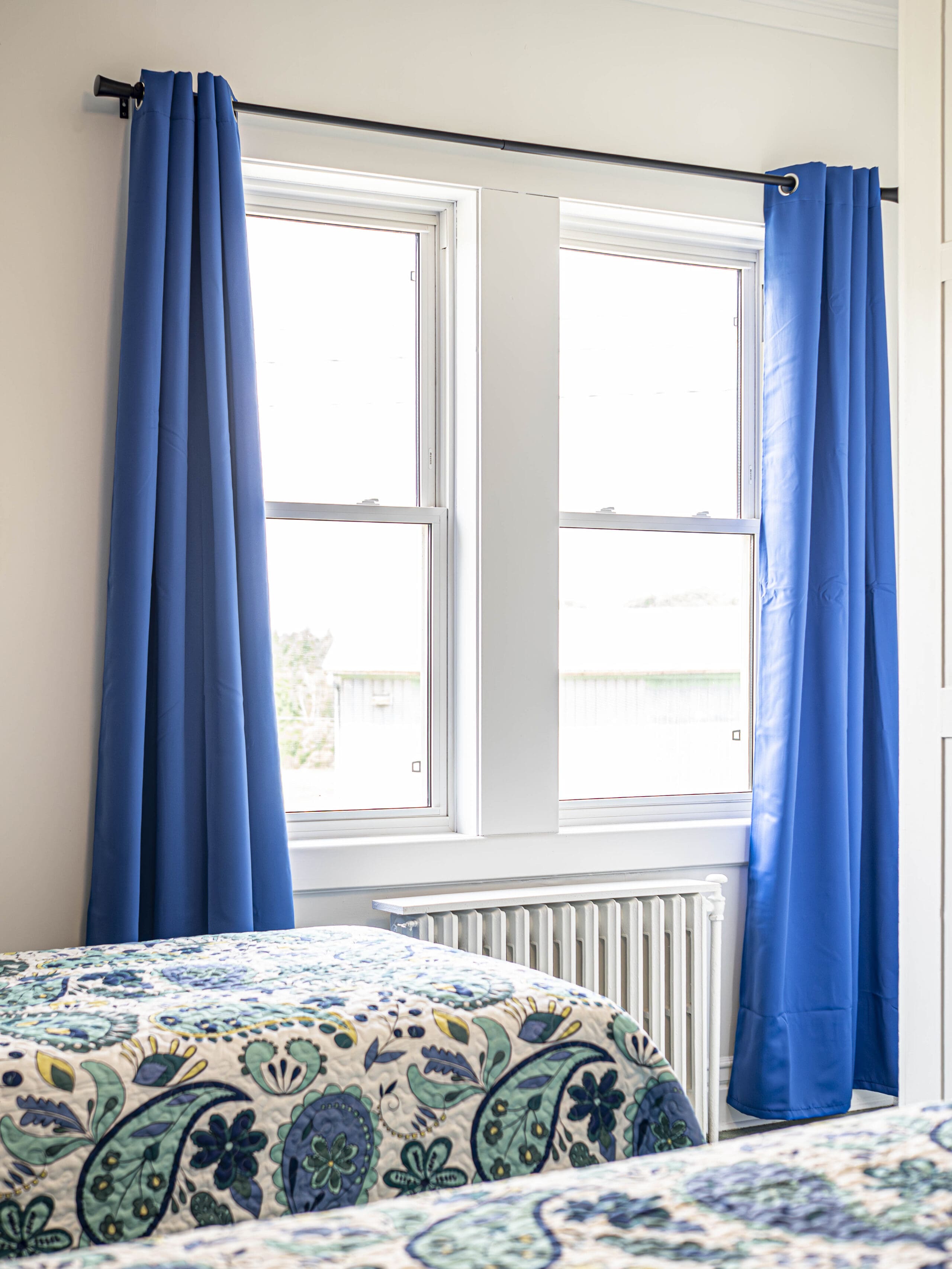 Chambre lumineuse avec deux lits jumeaux dotés de couettes à motifs floraux, de murs blancs, de deux grandes fenêtres et de longs rideaux bleus laissant passer la lumière du jour. Un radiateur blanc se trouve sous les fenêtres.