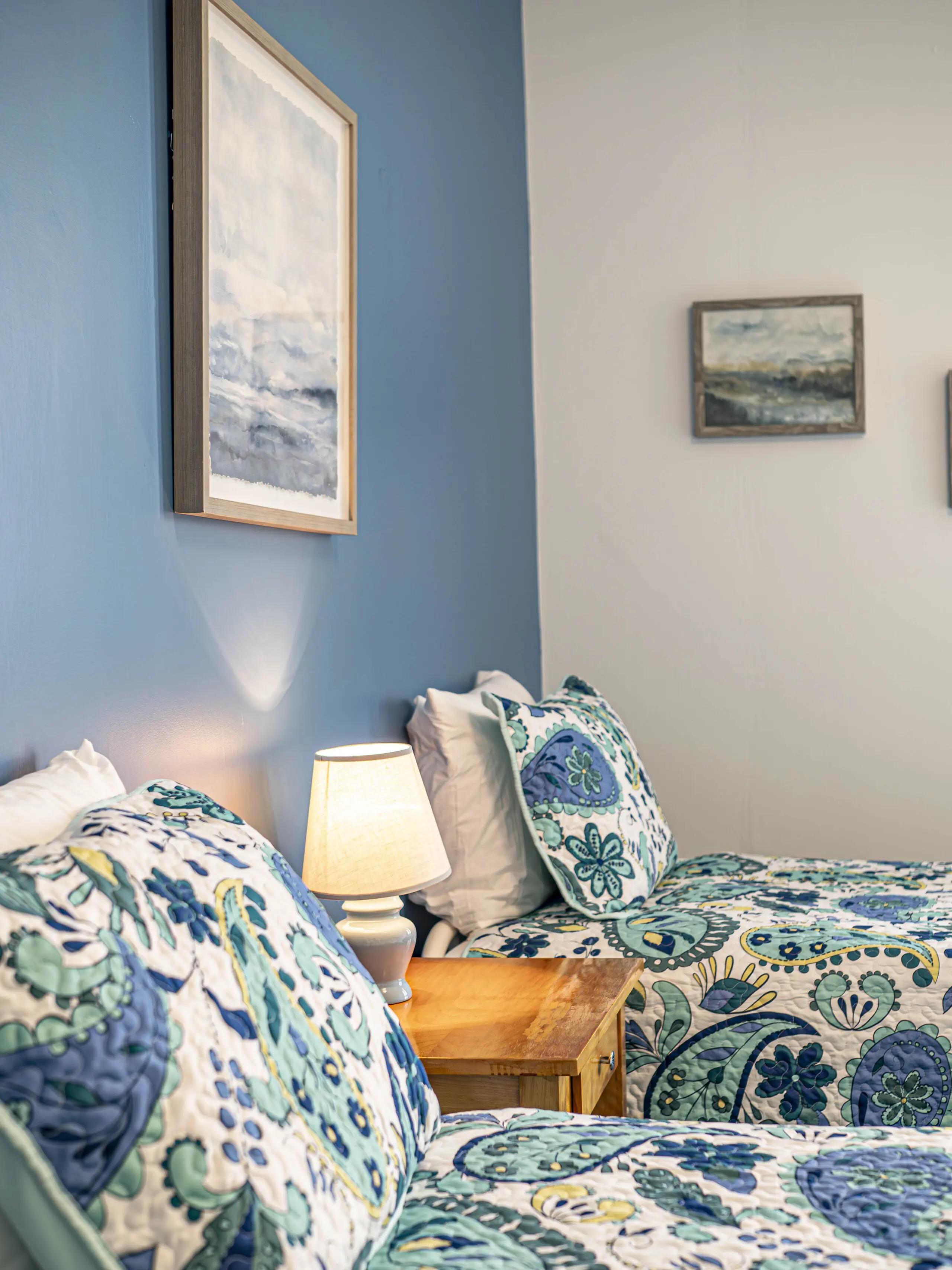 Deux lits jumeaux avec une literie colorée à fleurs bleues et vertes sont posés contre un mur d'accent bleu, avec une petite table de nuit en bois et une lampe allumée entre les deux. Des œuvres d'art encadrées sont suspendues au-dessus des lits.