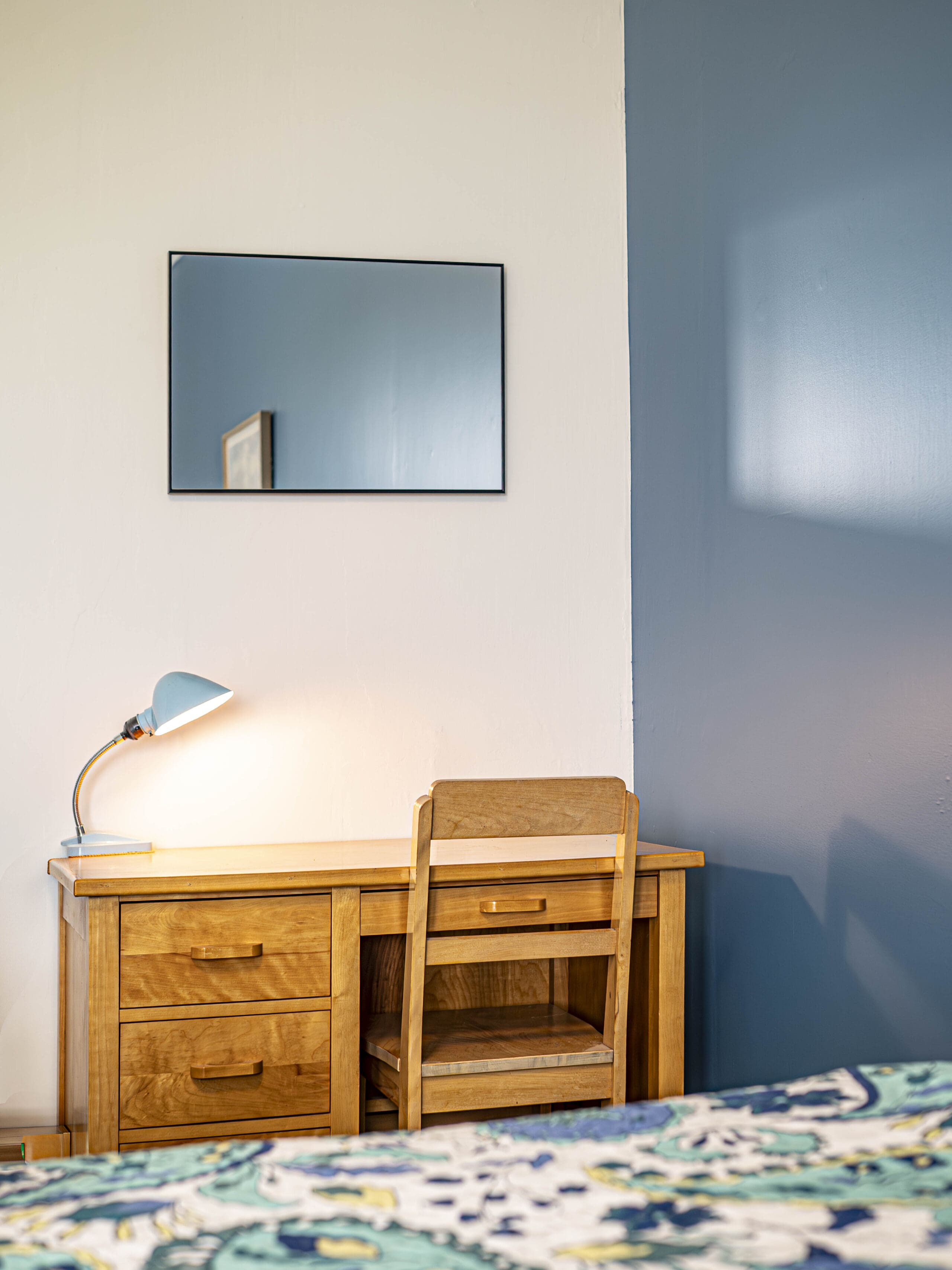 Un bureau en bois avec une chaise assortie, une lampe de bureau bleue et un petit miroir rectangulaire se trouvent sur un mur bicolore, en partie blanc et en partie bleu. Le bord d'un couvre-lit à motifs est visible au premier plan.