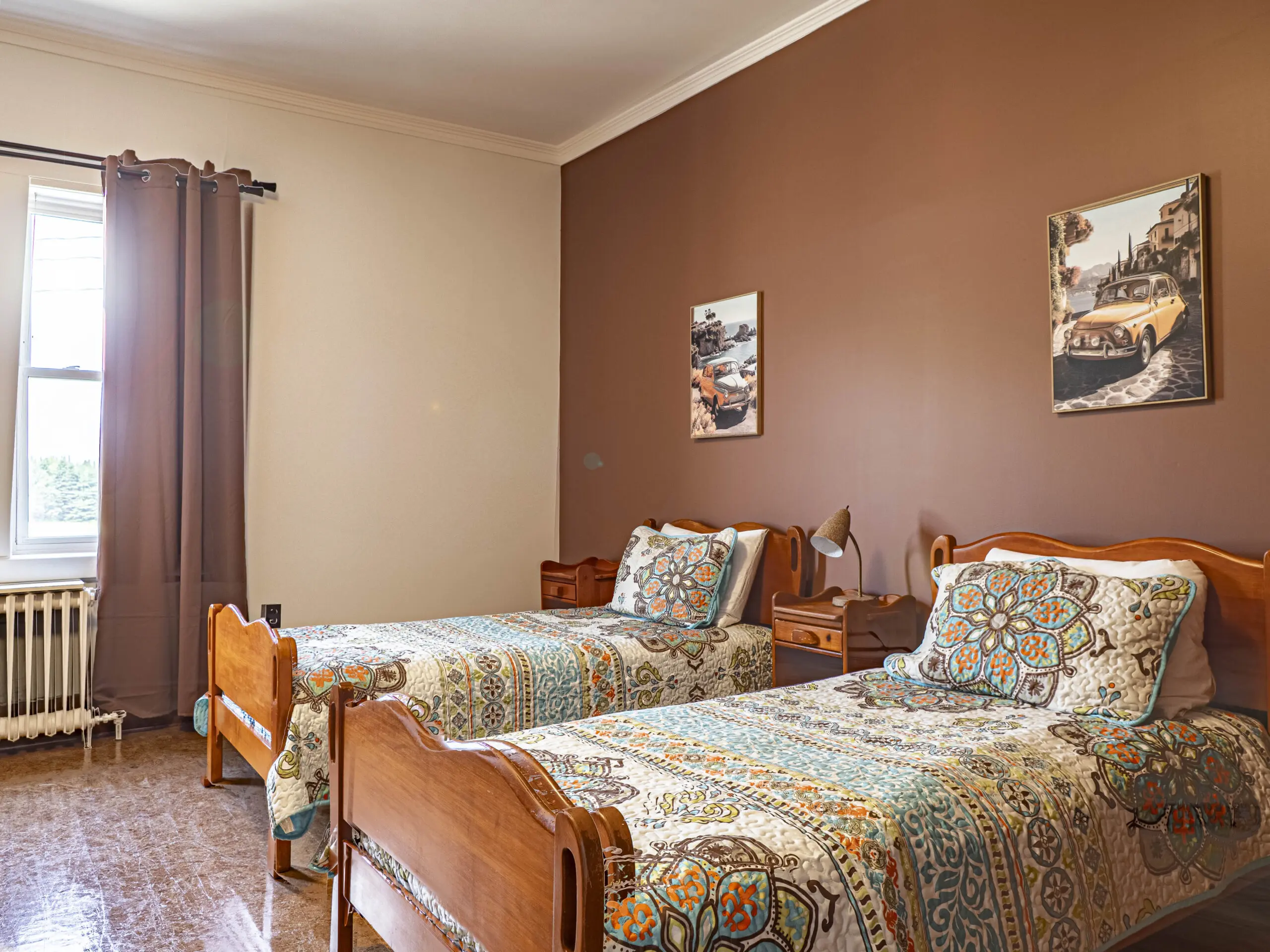 Une chambre confortable avec deux lits jumeaux en bois, des couvre-lits à motifs, un mur d'accent marron, deux tableaux sur le thème des voitures anciennes, une fenêtre avec des rideaux marron et un radiateur sous la fenêtre.