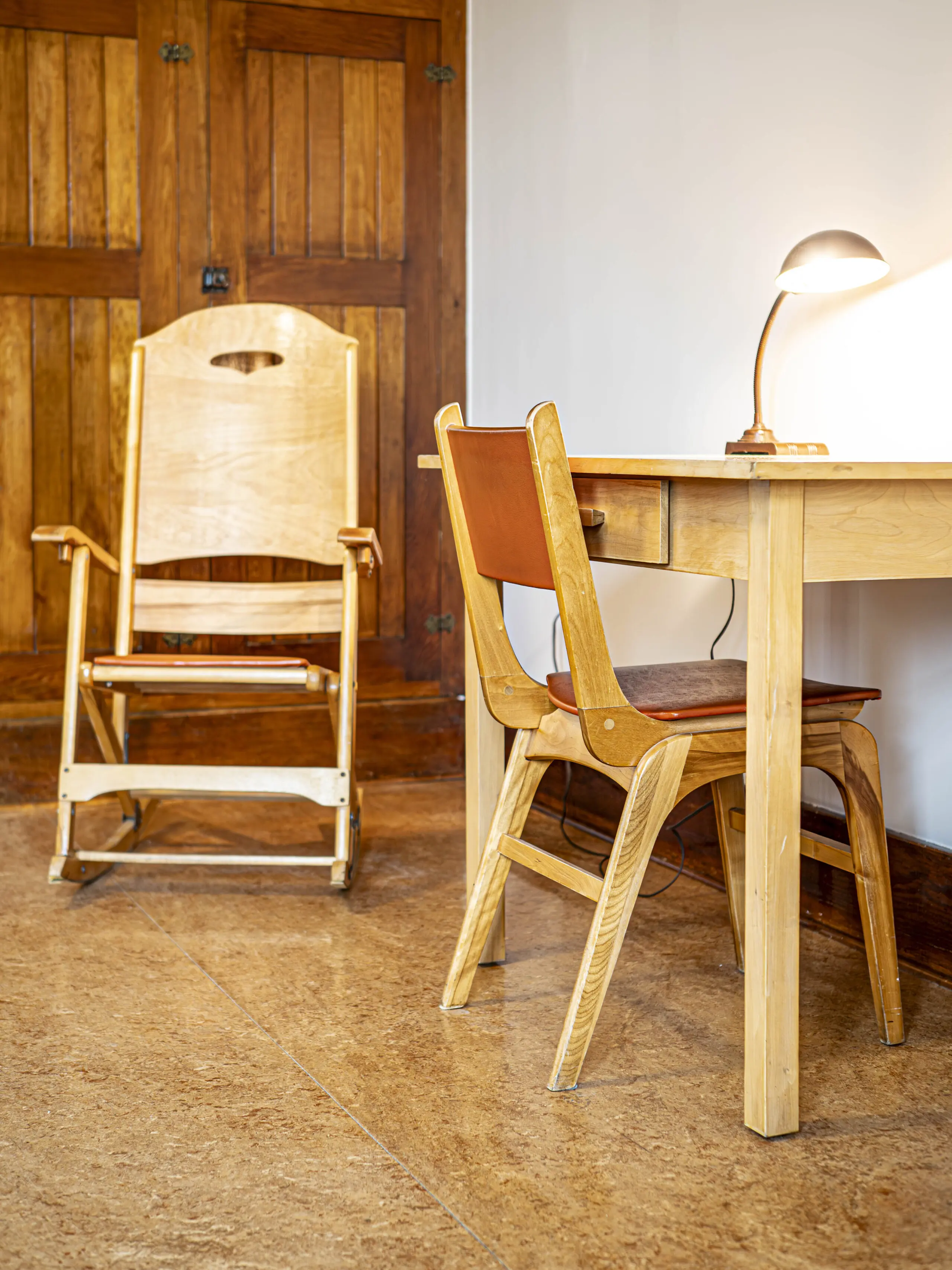 Un bureau en bois avec une chaise moderne et une petite lampe de bureau se trouve à côté d'une chaise à bascule en bois, le tout sur un sol en liège avec des portes en bois en arrière-plan.