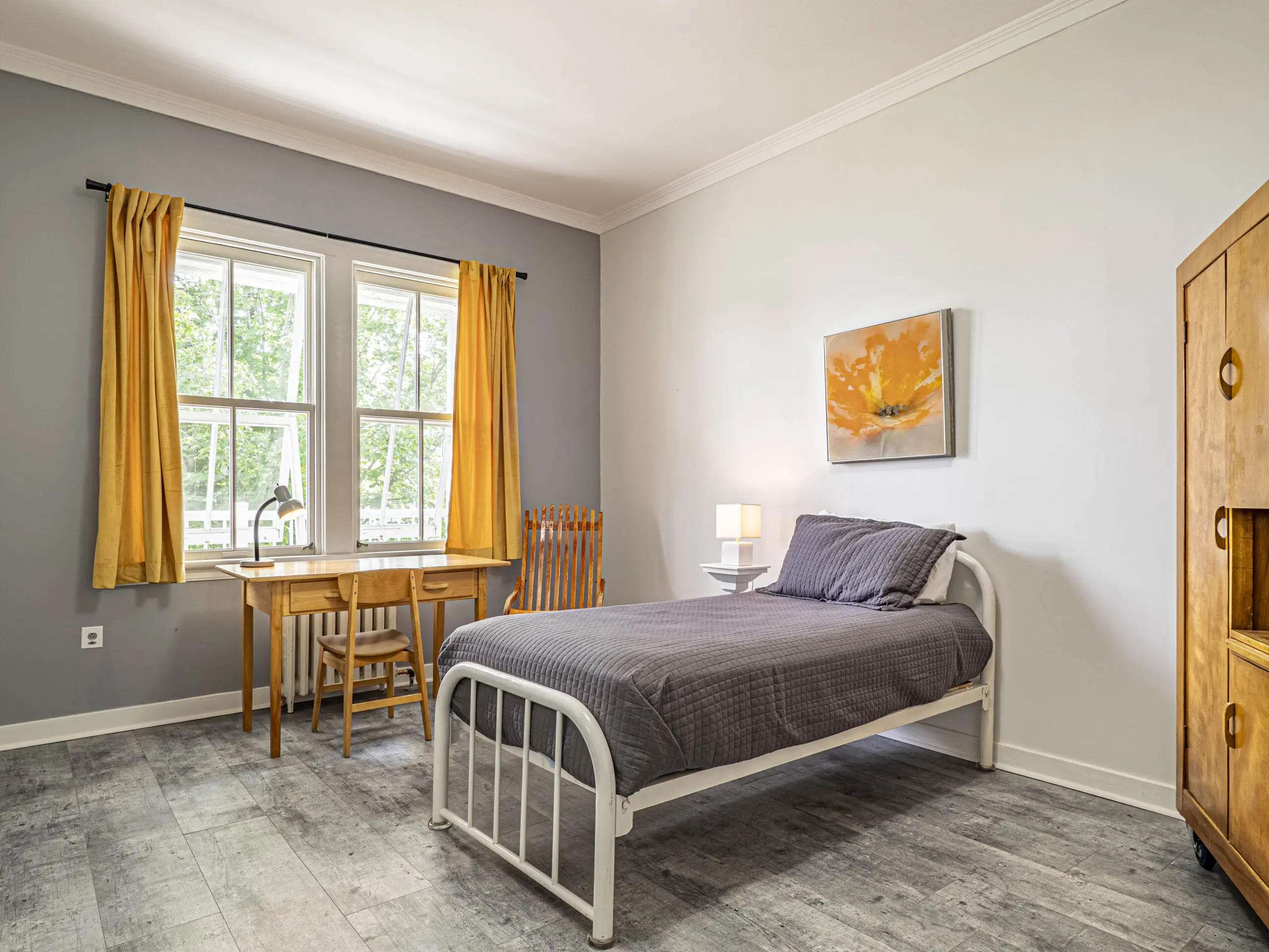 Une chambre lumineuse avec un lit simple, une literie grise, une armoire en bois, un bureau près de la fenêtre avec des rideaux jaunes, une chaise en bois et une table de nuit avec une lampe. Un tableau floral est accroché au-dessus du lit.