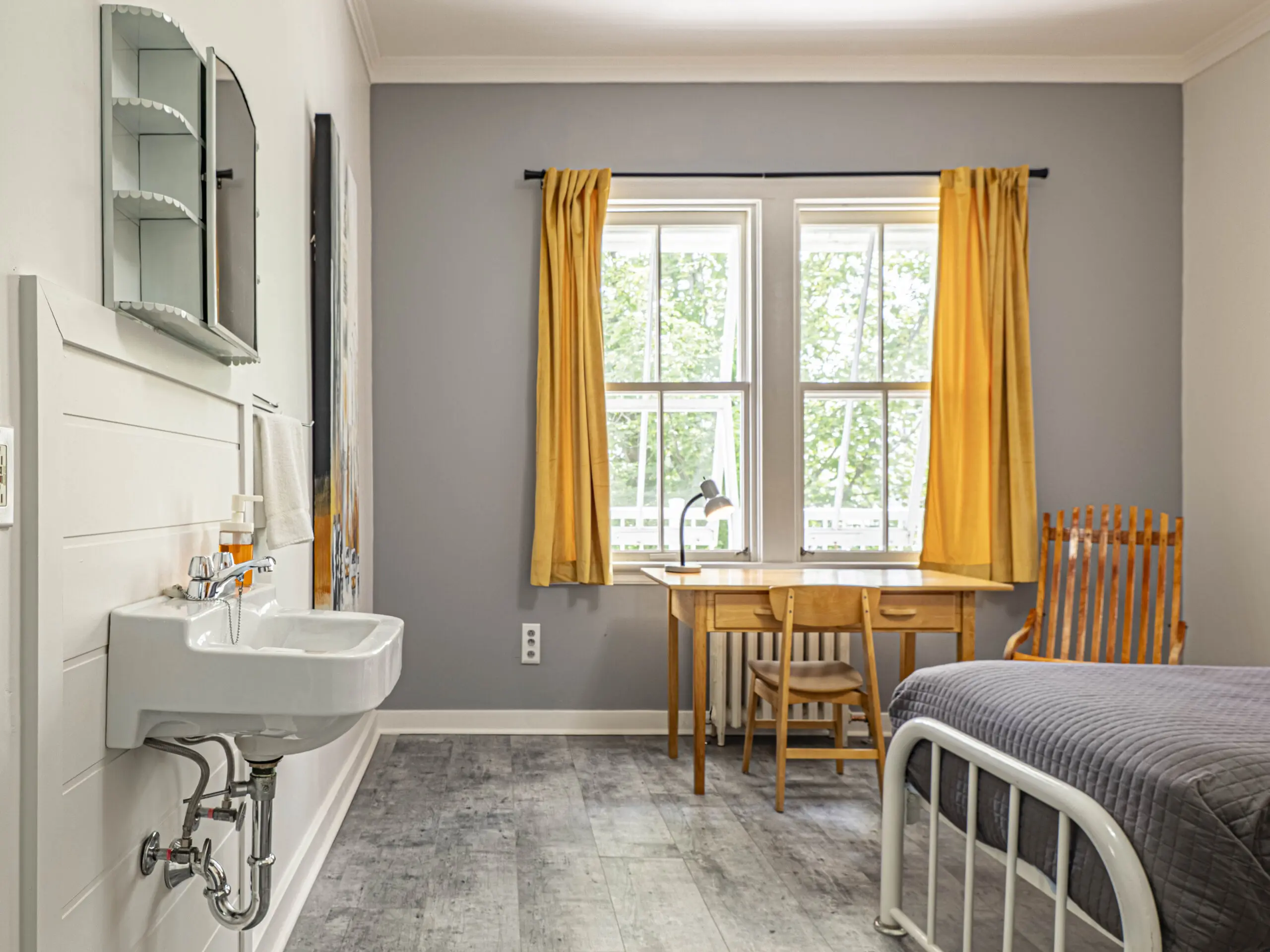 Une chambre bien rangée avec des murs gris, un lit avec un cadre gris, une chaise en bois et un bureau près de deux grandes fenêtres avec des rideaux jaunes, et un évier blanc fixé au mur avec un miroir à gauche.