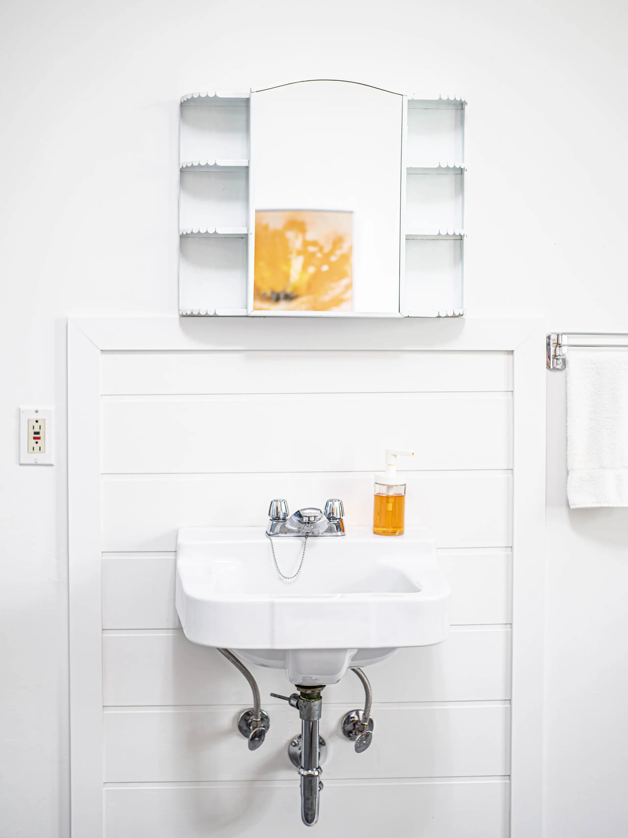 Un petit lavabo blanc fixé au mur avec un robinet chromé se trouve sous une armoire à pharmacie à miroir. Un distributeur de savon orange est posé sur le lavabo et une serviette est suspendue à droite. Les murs et la décoration sont entièrement blancs.