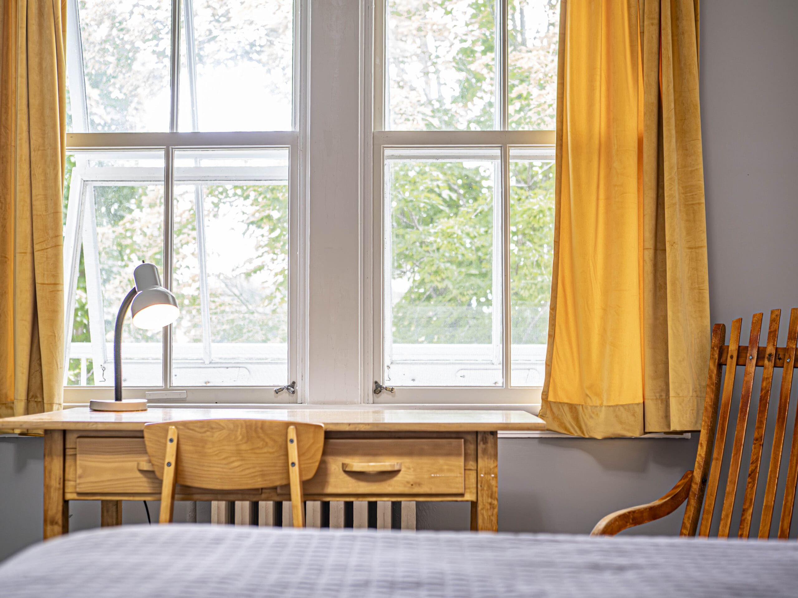 Un bureau en bois avec une chaise et une lampe se trouve devant une grande fenêtre aux rideaux jaunes. Un rocking-chair en bois se trouve à droite et un lit avec une couverture grise se trouve au premier plan. Des arbres sont visibles à l'extérieur de la fenêtre.