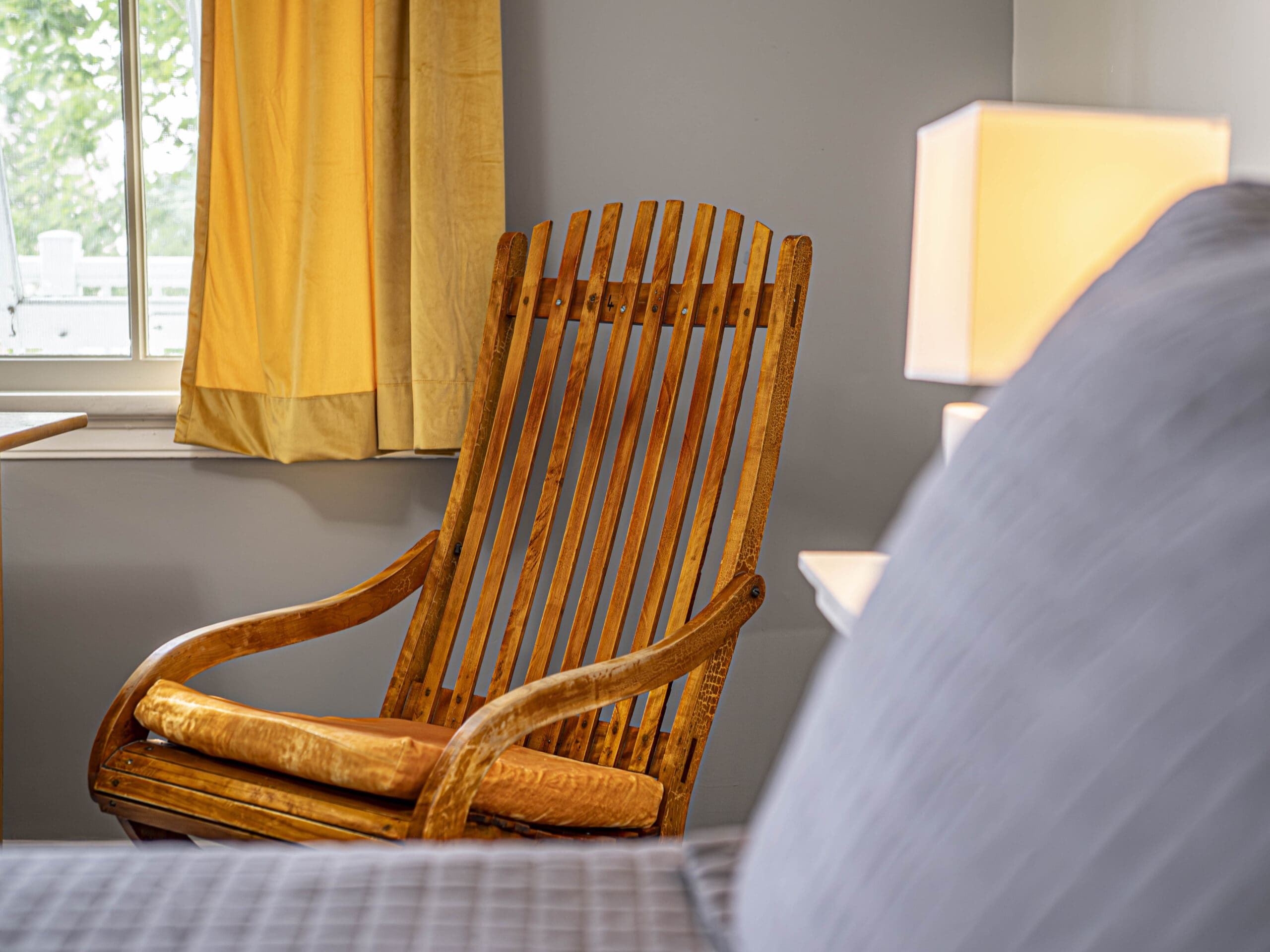 Une chaise à bascule en bois avec un coussin jaune est assise près d'une fenêtre avec des rideaux jaunes, à côté d'une lampe rougeoyante et d'un lit avec une couverture matelassée grise dans une chambre confortable.