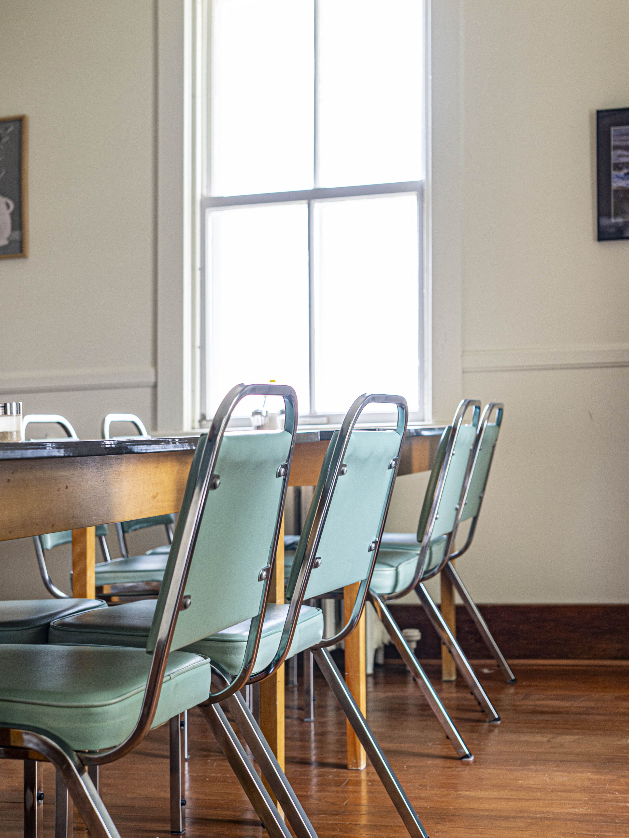 Une rangée de chaises vintage vert menthe à armature métallique est alignée le long d'une table en bois dans une pièce lumineuse dotée d'une grande fenêtre et d'un plancher en bois dur.