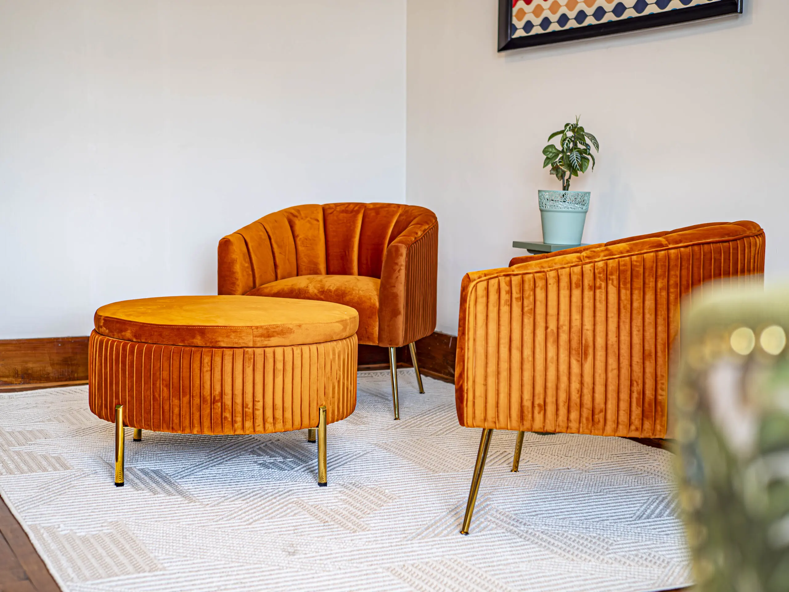 Deux chaises en velours orange et un ottoman rond assorti aux pieds dorés sont disposés sur un tapis clair dans une pièce moderne. Une petite table d'appoint avec une plante en pot est posée entre les chaises, et des œuvres d'art sont accrochées au mur blanc.