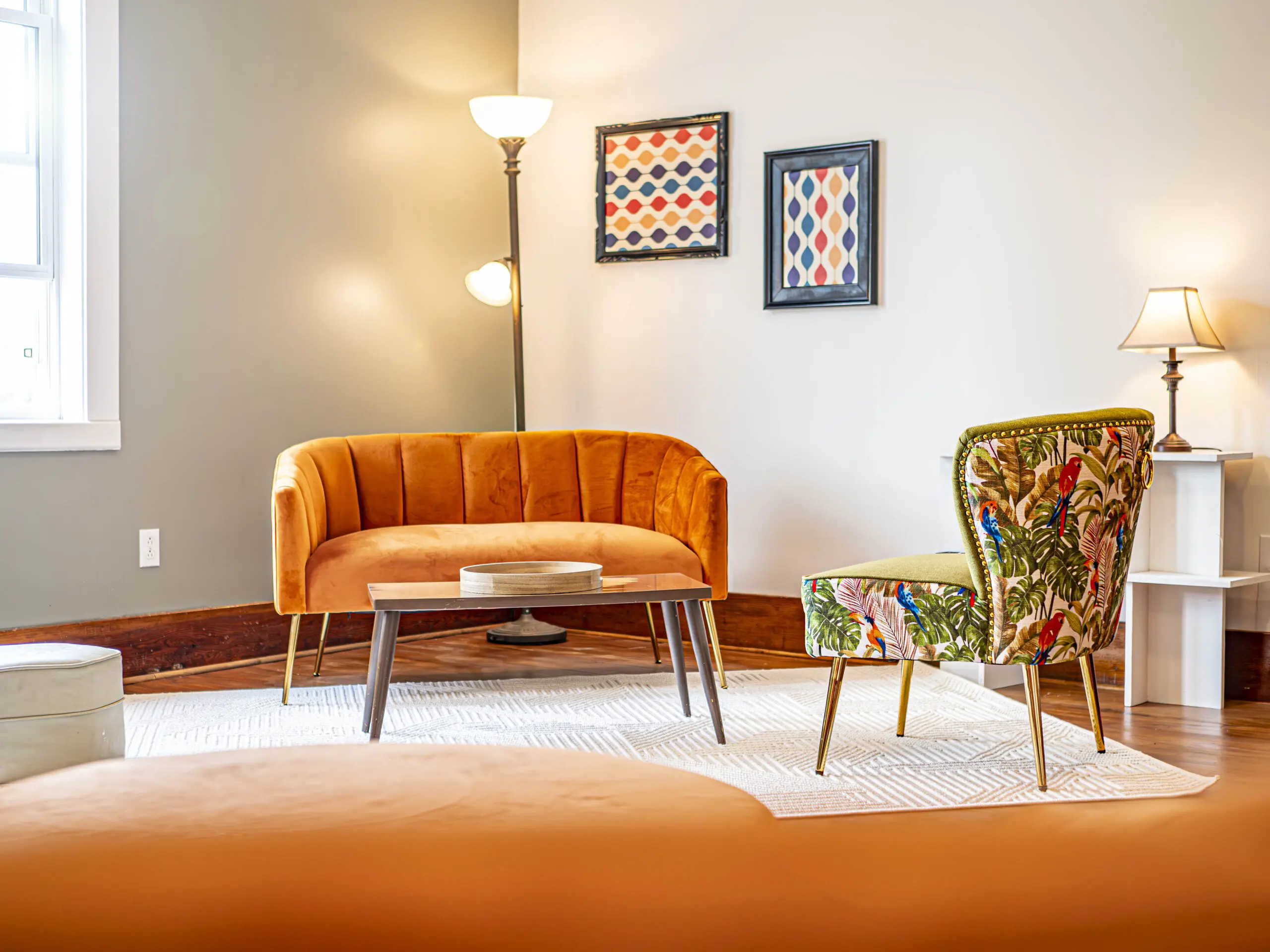 Un salon confortable avec une causeuse en velours orange, une chaise à motifs floraux, un lampadaire et une petite lampe de table. Deux œuvres d'art encadrées sont accrochées au mur au-dessus d'un tapis blanc et d'un plancher en bois clair.