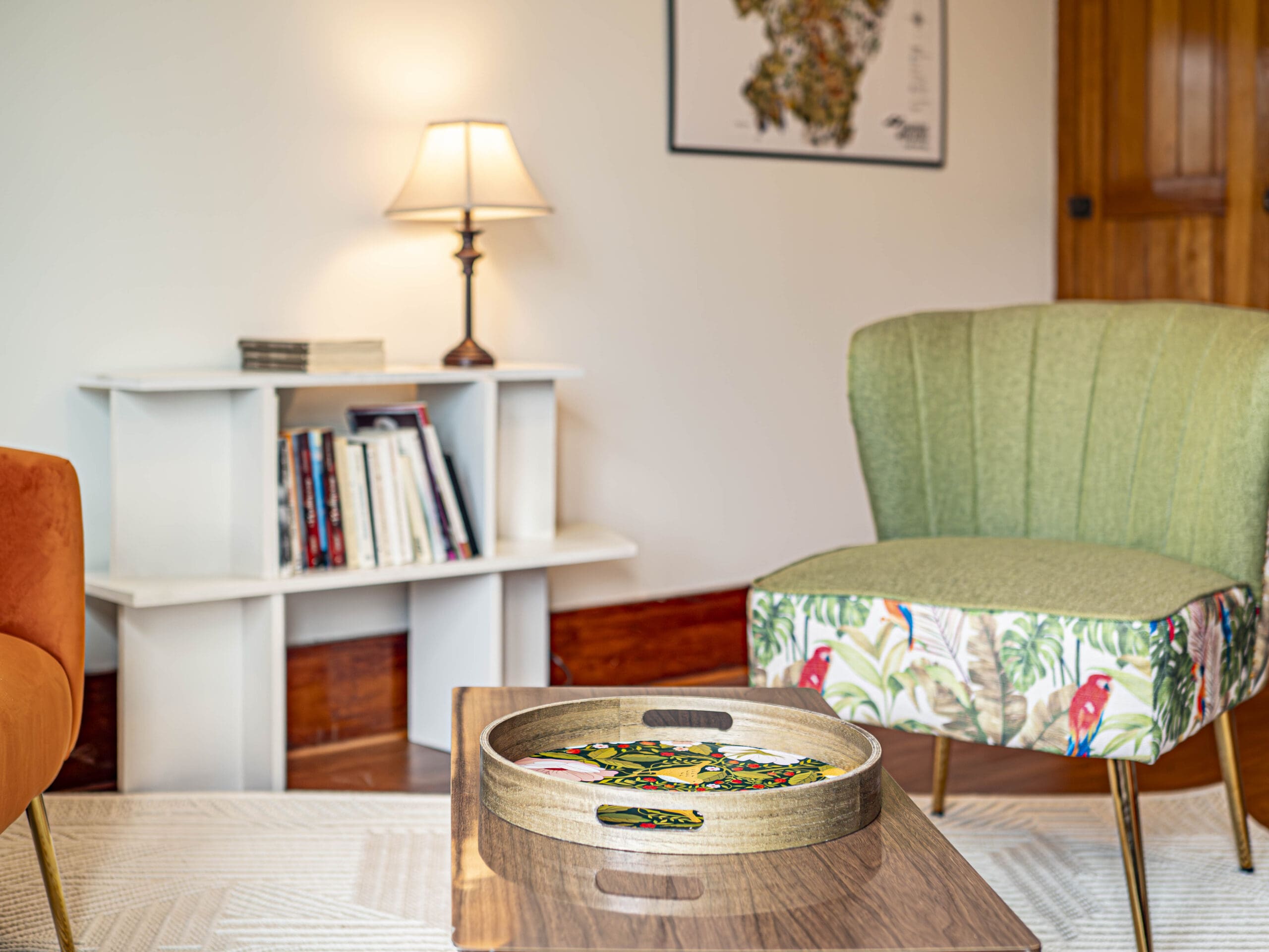 Un salon confortable avec un fauteuil vert, une table basse en bois contenant un plateau décoratif, une chaise orange, une petite étagère avec des livres, une lampe allumée et une carte encadrée au mur.