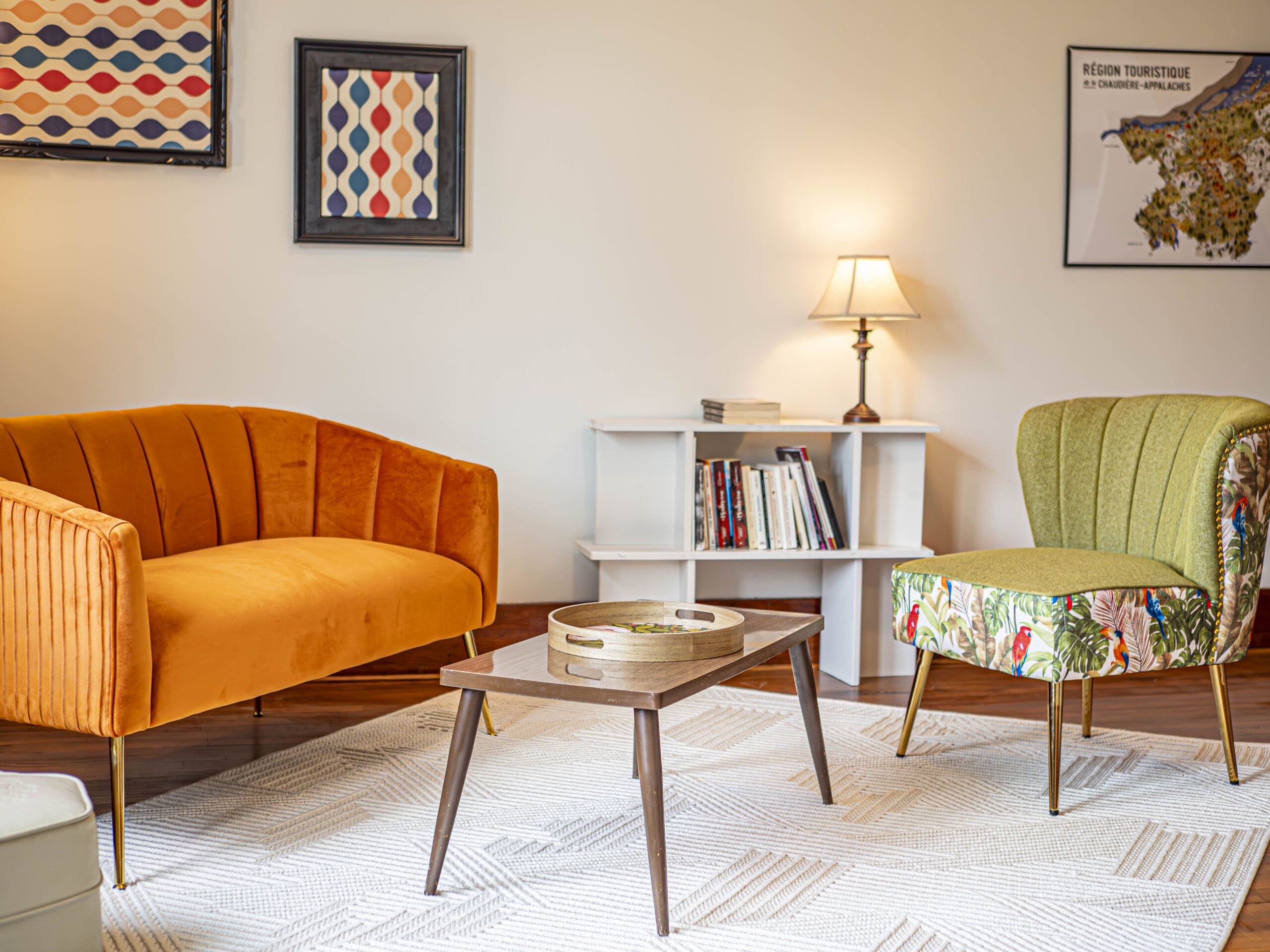 Un salon confortable avec un canapé en velours orange, une chaise verte à motifs floraux, une table basse en bois, une étagère blanche avec des livres, une lampe et des œuvres d'art encadrées sur des murs de couleur crème.