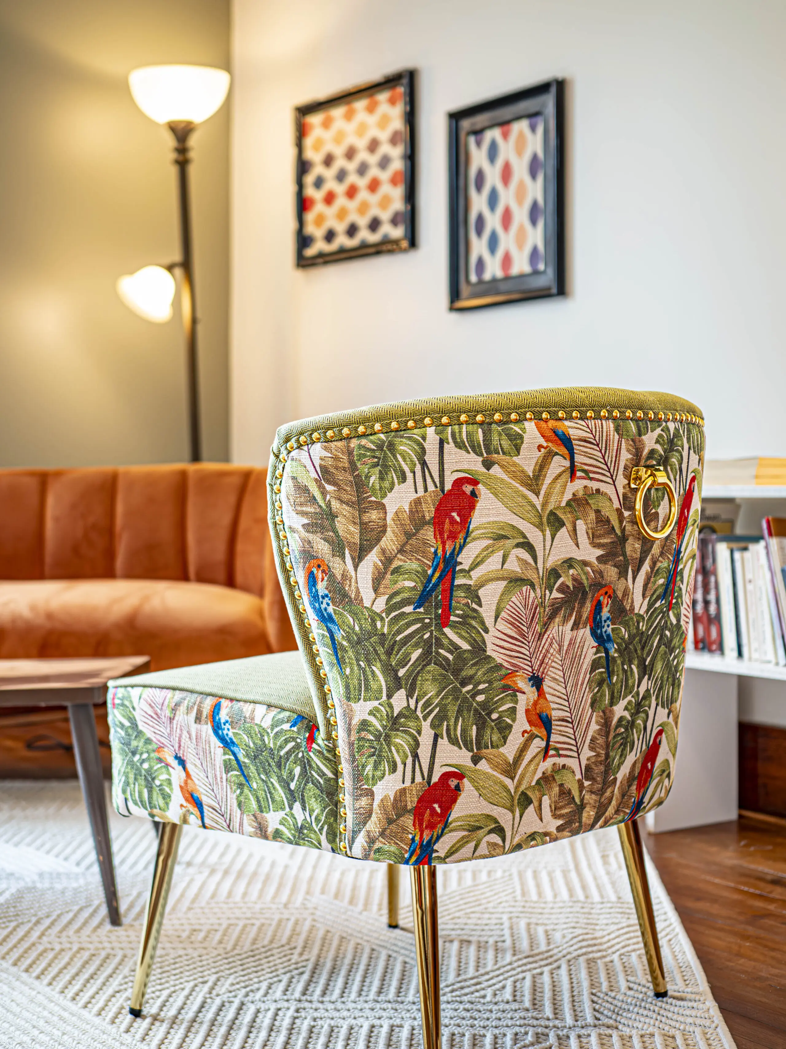 Un salon élégant avec une chaise à motifs représentant des perroquets colorés et des feuilles tropicales, un canapé orange en daim, une table basse en bois et des œuvres d'art encadrées au mur. Un grand lampadaire éclaire doucement l'espace.