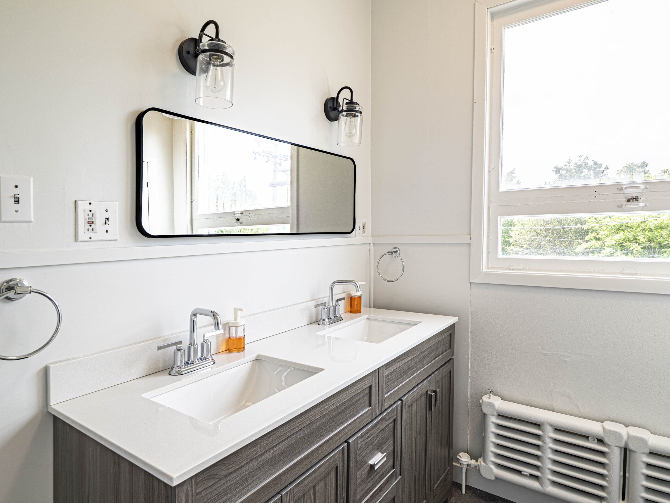 Salle de bains moderne avec deux lavabos, un grand miroir rectangulaire, des appliques noires, des distributeurs de savon, des anneaux porte-serviettes et une fenêtre laissant entrer la lumière naturelle. Le meuble-lavabo est en bois foncé et le plan de travail est blanc.