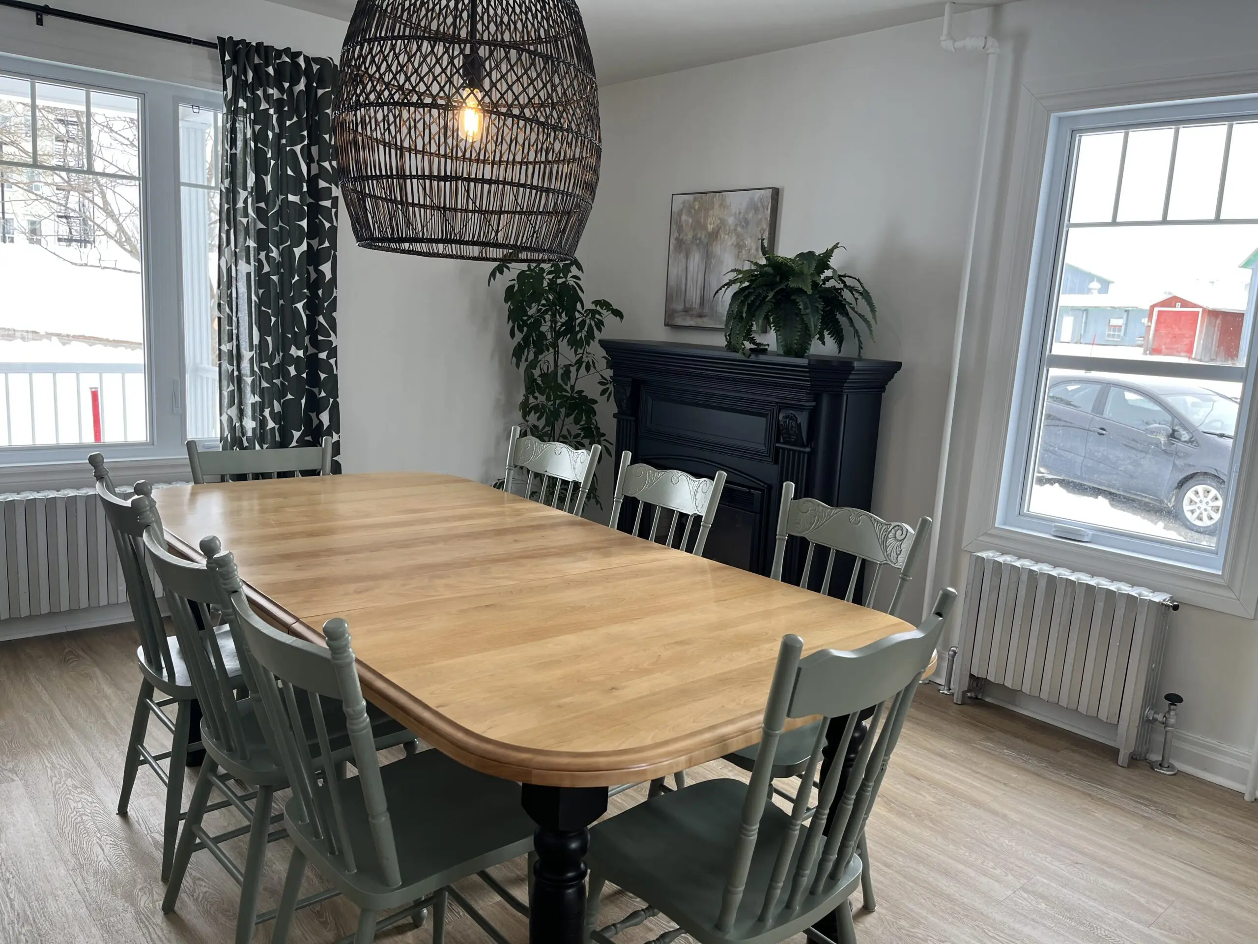 Une salle à manger avec une table en bois clair entourée de chaises vertes, une cheminée noire, des plantes en pot, des rideaux à motifs, de grandes fenêtres et une suspension tissée suspendue au-dessus de la table.