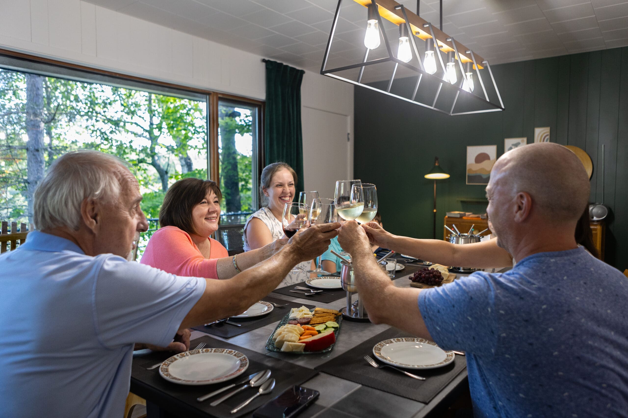 Quatre adultes trinquant joyeusement avec des verres à vin à une table dans une pièce bien éclairée avec une fenêtre donnant sur la verdure à l'extérieur.