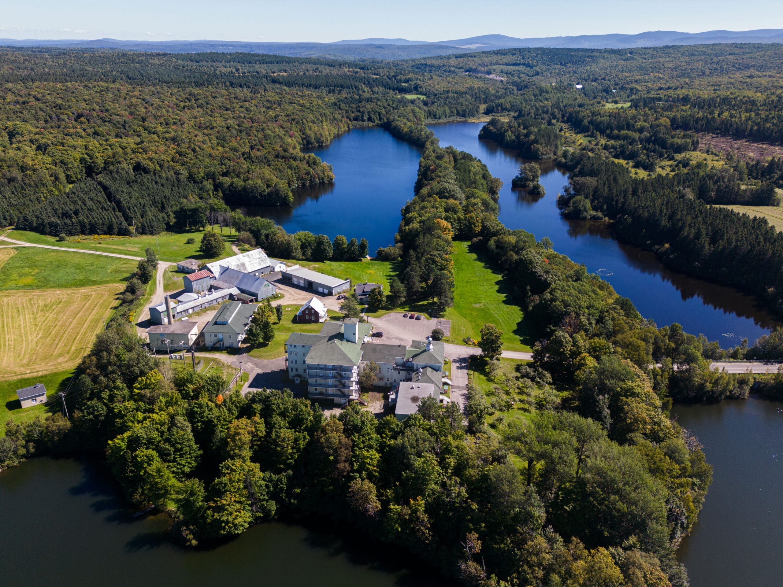 Vue aérienne d'un grand domaine avec plusieurs bâtiments à côté d'un lac pittoresque entouré de forêts luxuriantes sous un ciel clair.