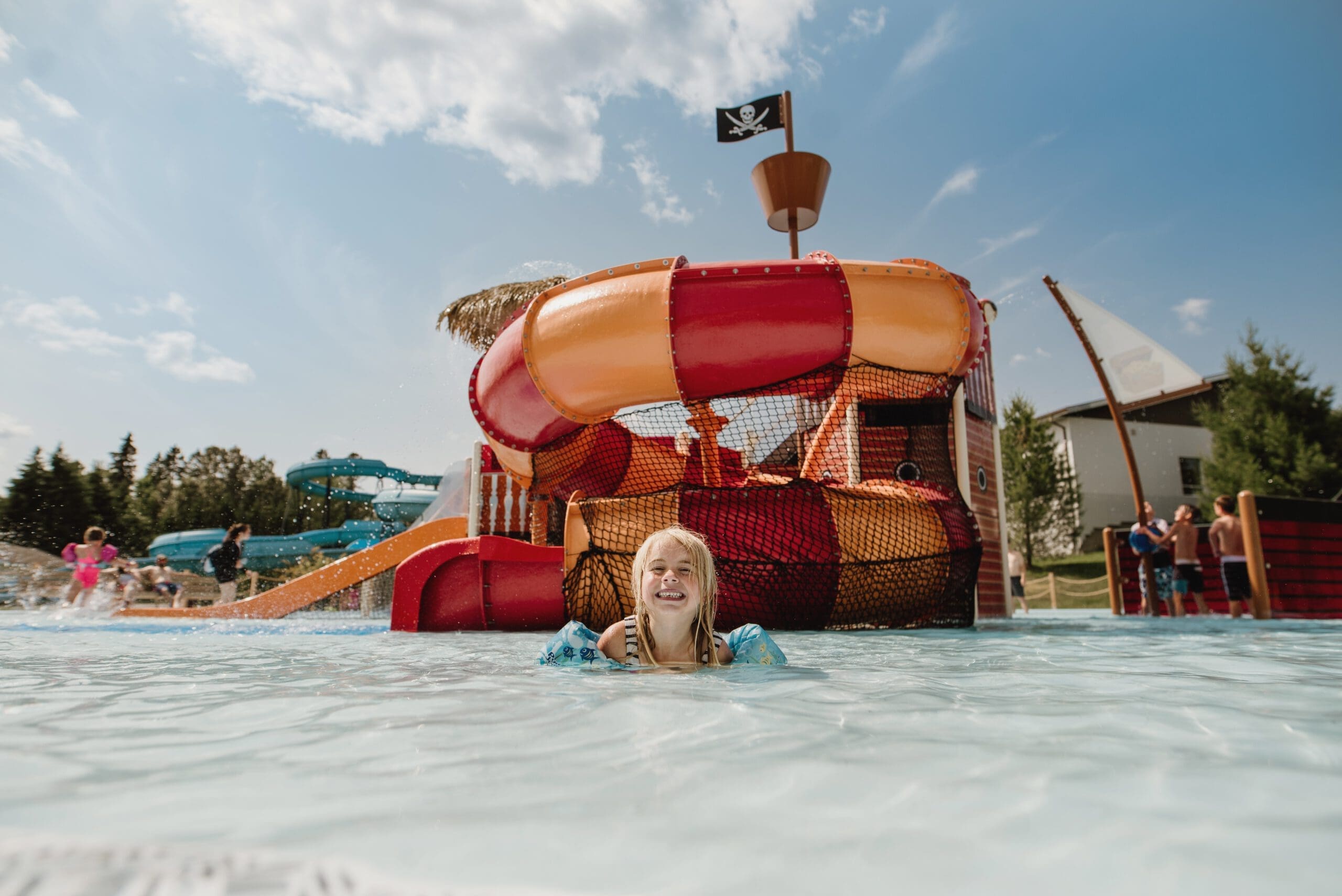 Un enfant joyeux aux cheveux blonds souriant dans une piscine, avec en arrière-plan un grand toboggan gonflable sur le thème d'un bateau pirate sous un ciel ensoleillé.