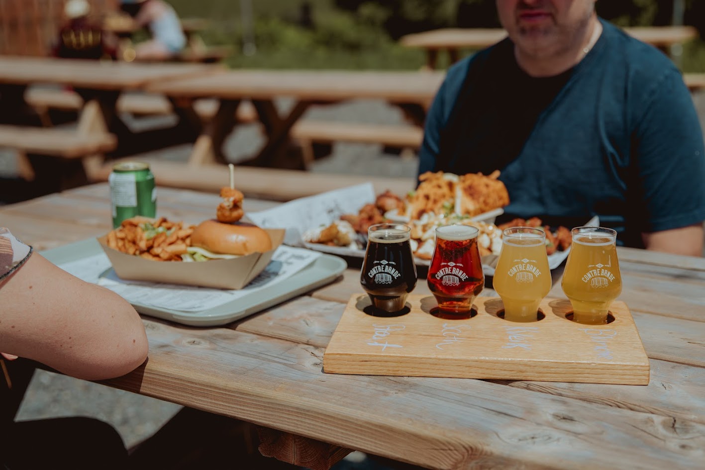 Une scène de repas en plein air avec une table en bois contenant une série de bières variées, un hamburger, des frites et d'autres entrées, avec des personnes partiellement visibles en arrière-plan.