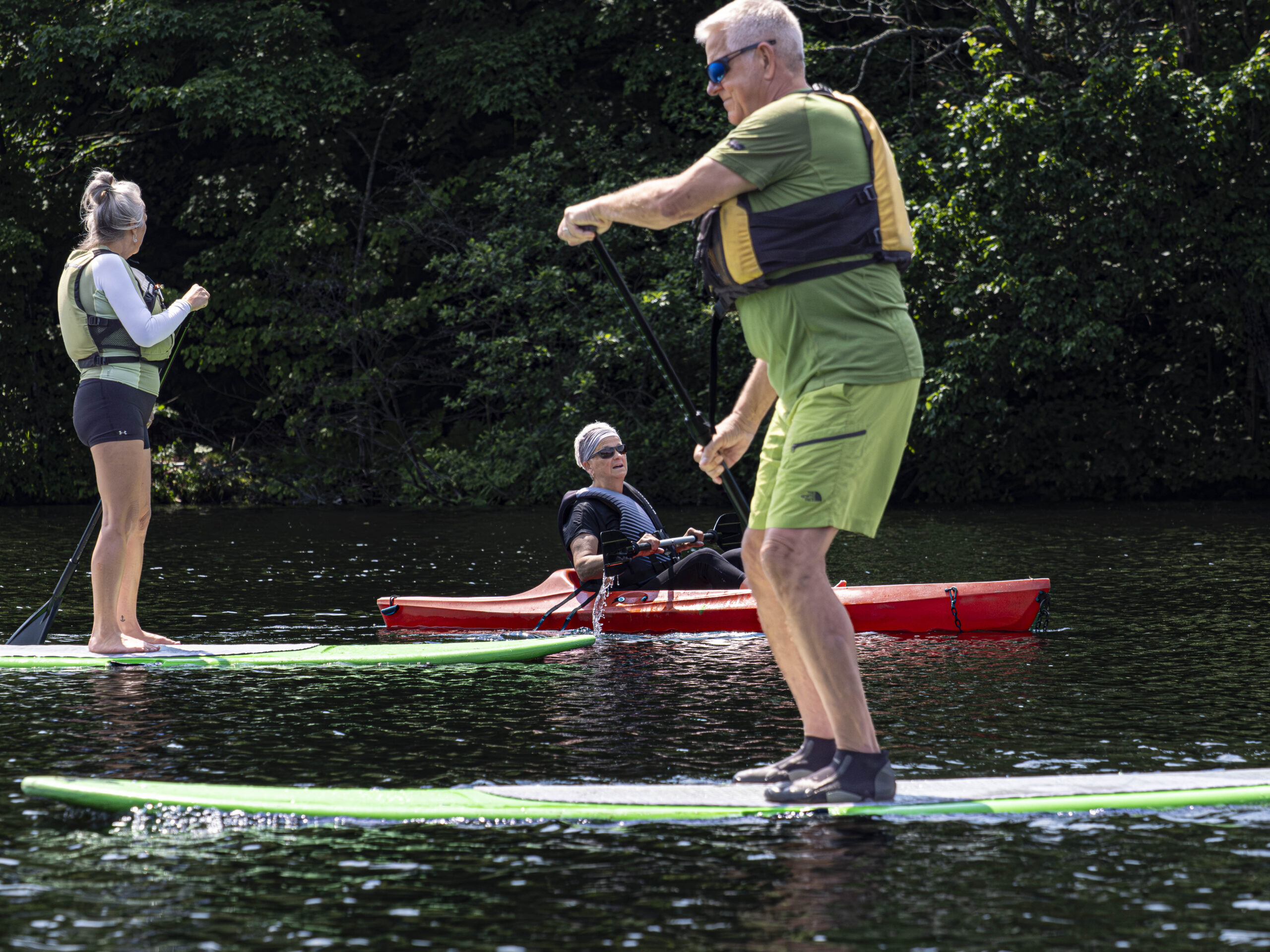 Deux personnes sont debout sur des planches à pagayer tandis qu'une autre personne dans un kayak rouge pagaie à proximité sur un lac calme, entouré d'arbres et de verdure par une journée ensoleillée.