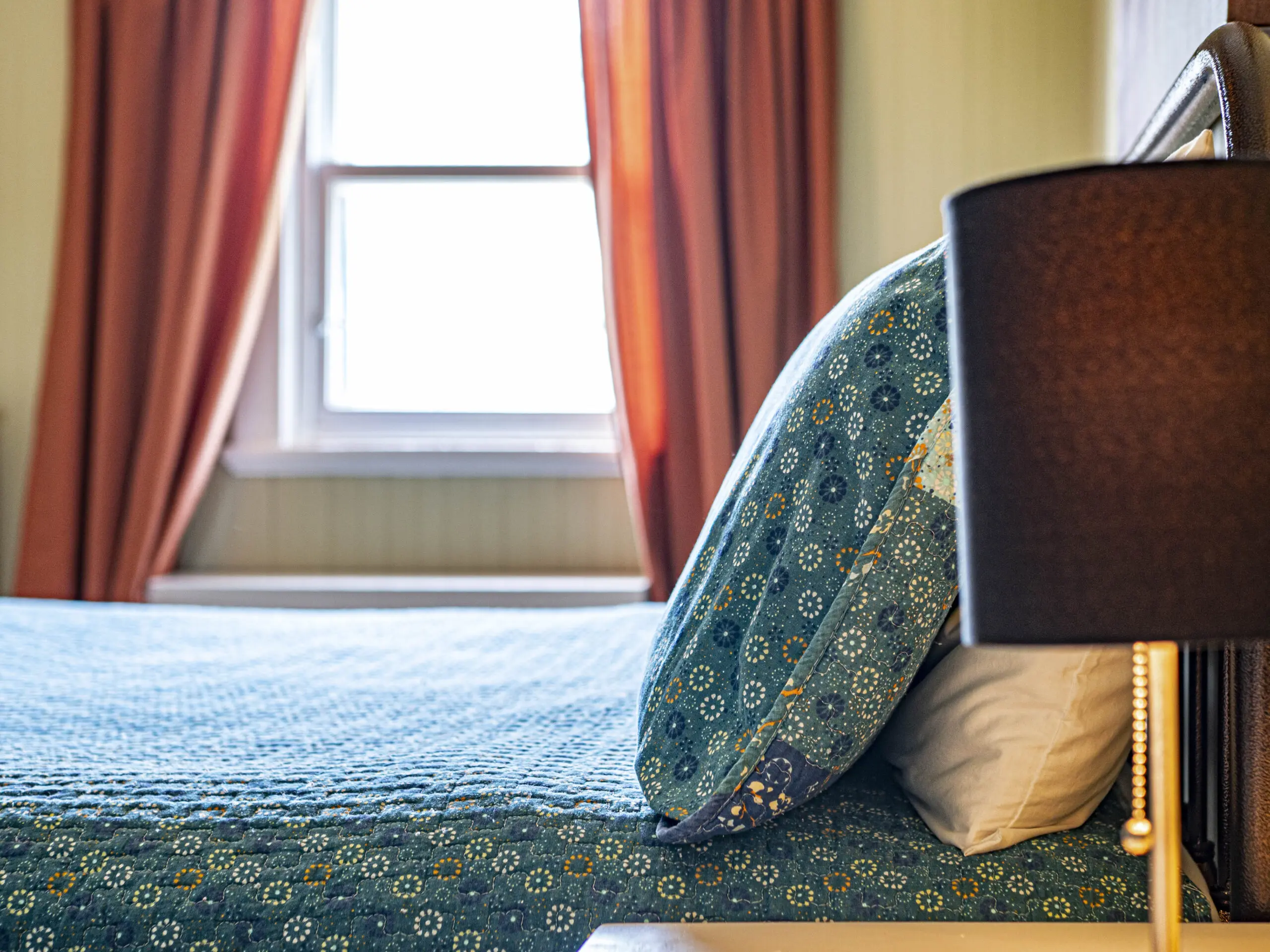 Une chambre à coucher confortable avec un lit soigneusement fait avec des draps à motifs bleus, un oreiller, un abat-jour noir sur une table de nuit, et la lumière du soleil qui traverse une fenêtre avec des rideaux orange en arrière-plan.