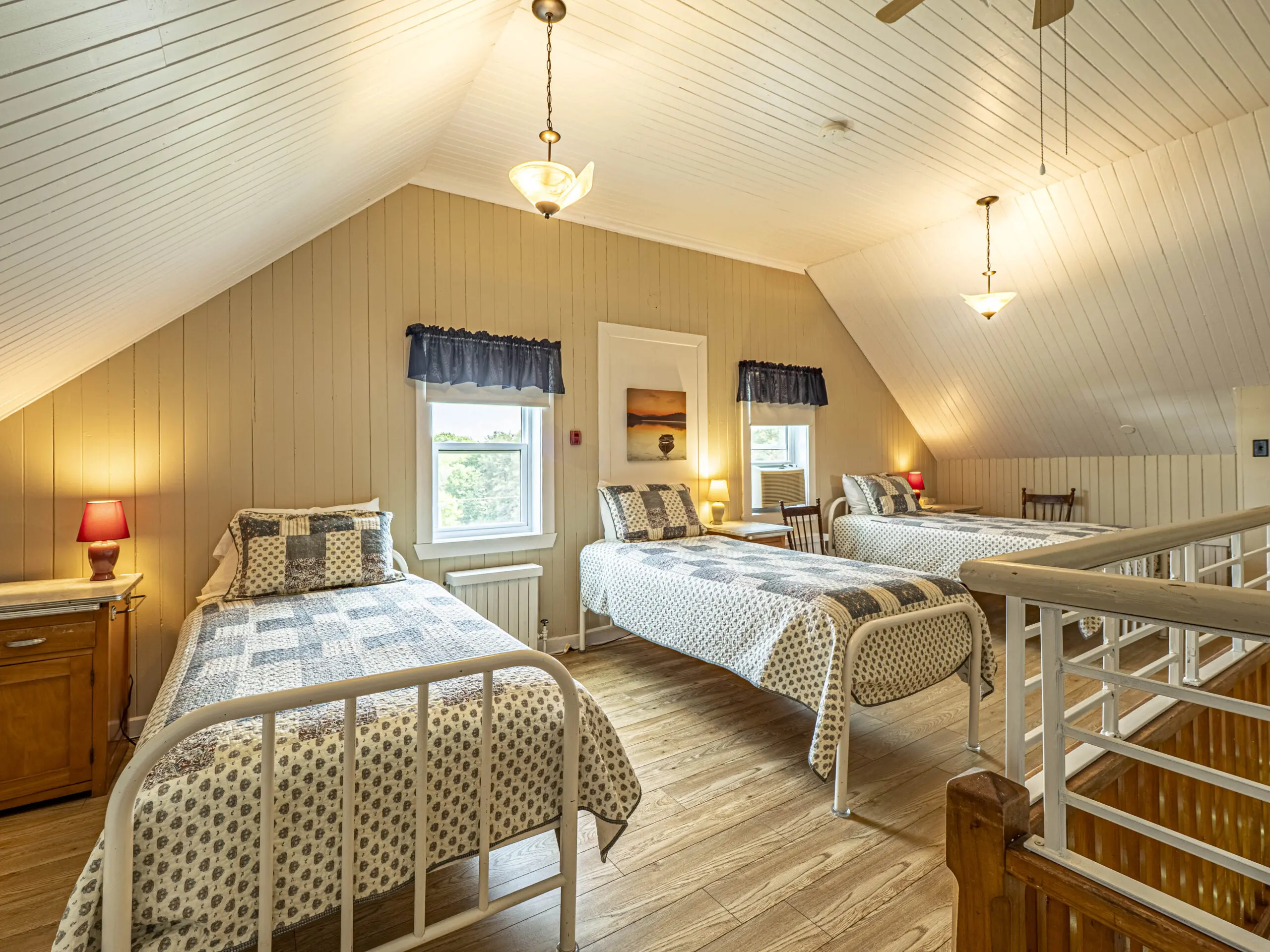 Une chambre mansardée confortable avec trois lits simples, une literie à motifs, des planchers en bois et des plafonds inclinés. Deux fenêtres avec des rideaux bleu marine laissent entrer la lumière naturelle. Des lampes et un éclairage chaleureux créent une atmosphère accueillante.