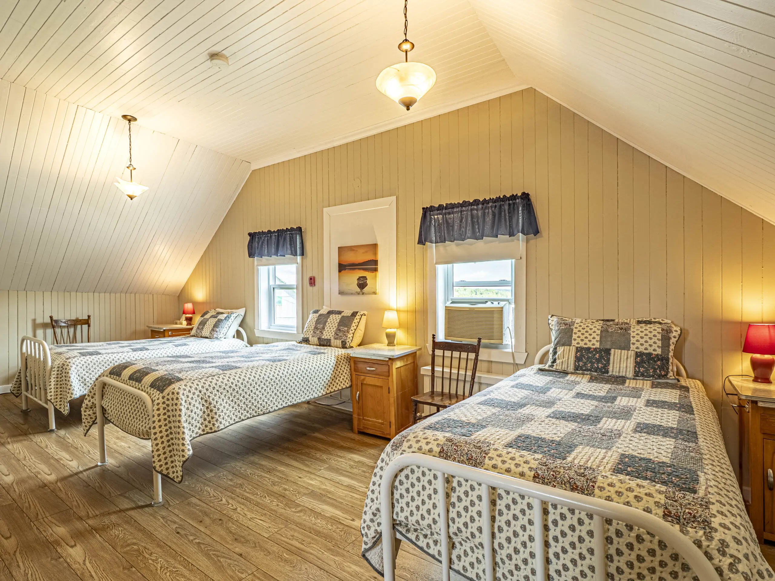 Chambre mansardée confortable avec trois lits à armature métallique, chacun avec des couettes en patchwork, des tables de nuit en bois avec des lampes, une chaise en bois, deux fenêtres avec des rideaux bleus, un plafond incliné à panneaux de bois et un éclairage chaleureux.
