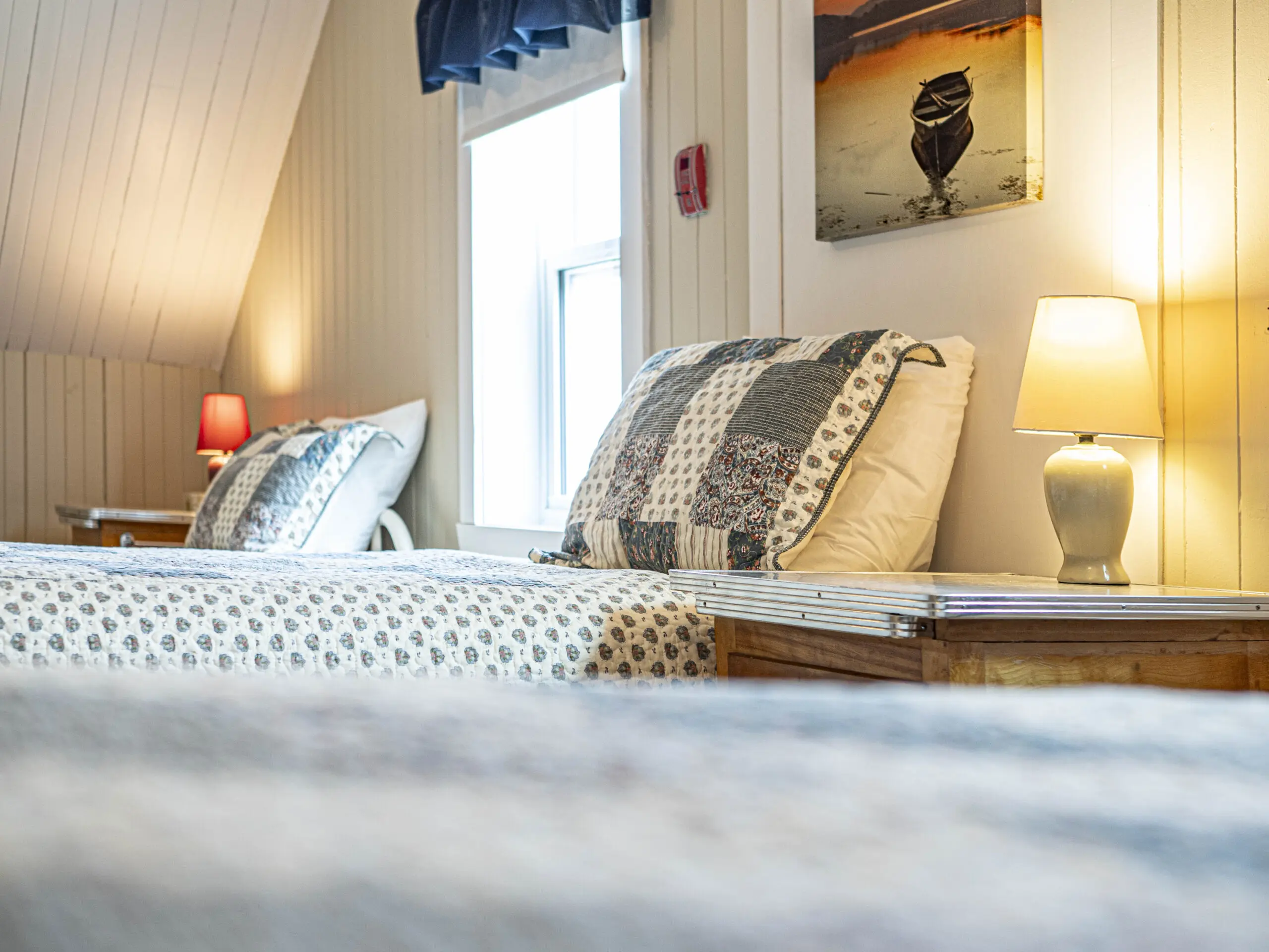Une chambre confortable avec deux lits recouverts d'édredons à motifs, des tables de chevet avec des lampes allumées, une fenêtre avec des rideaux bleus et un tableau encadré au mur. La pièce est éclairée par une lumière douce et le plafond est incliné.