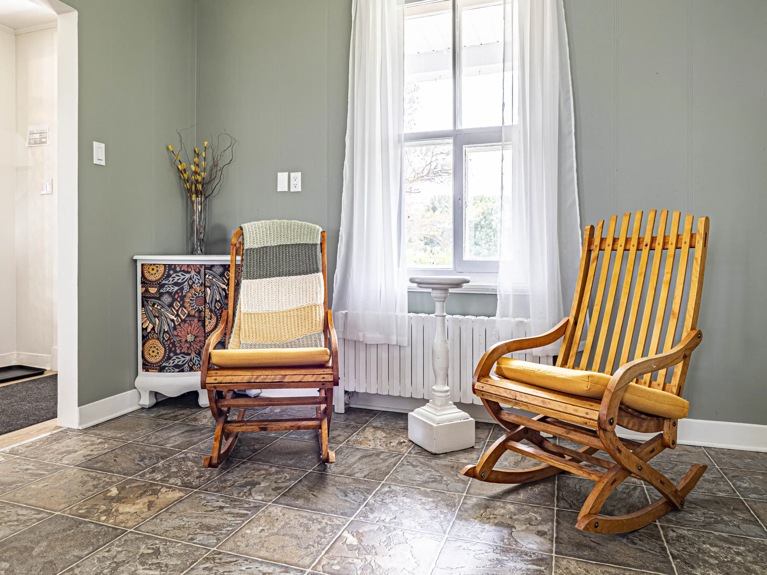 Deux fauteuils à bascule en bois sont posés sur un sol carrelé, près d'une fenêtre aux rideaux blancs, dans une pièce confortable et ensoleillée. Une petite armoire au design coloré et un vase décoratif sont également visibles.