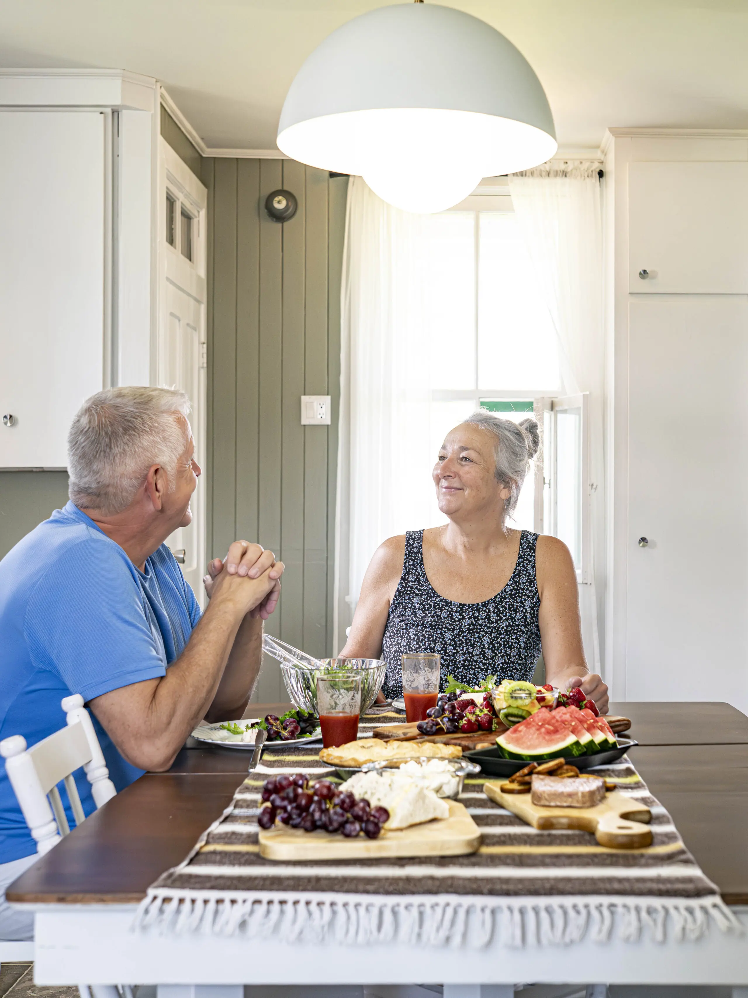 Un homme et une femme âgés sont assis à une table à manger, se souriant l'un à l'autre. La table est garnie de fruits, de fromage, de crackers et de boissons. La pièce est baignée de lumière naturelle, avec des armoires blanches et une lampe suspendue.