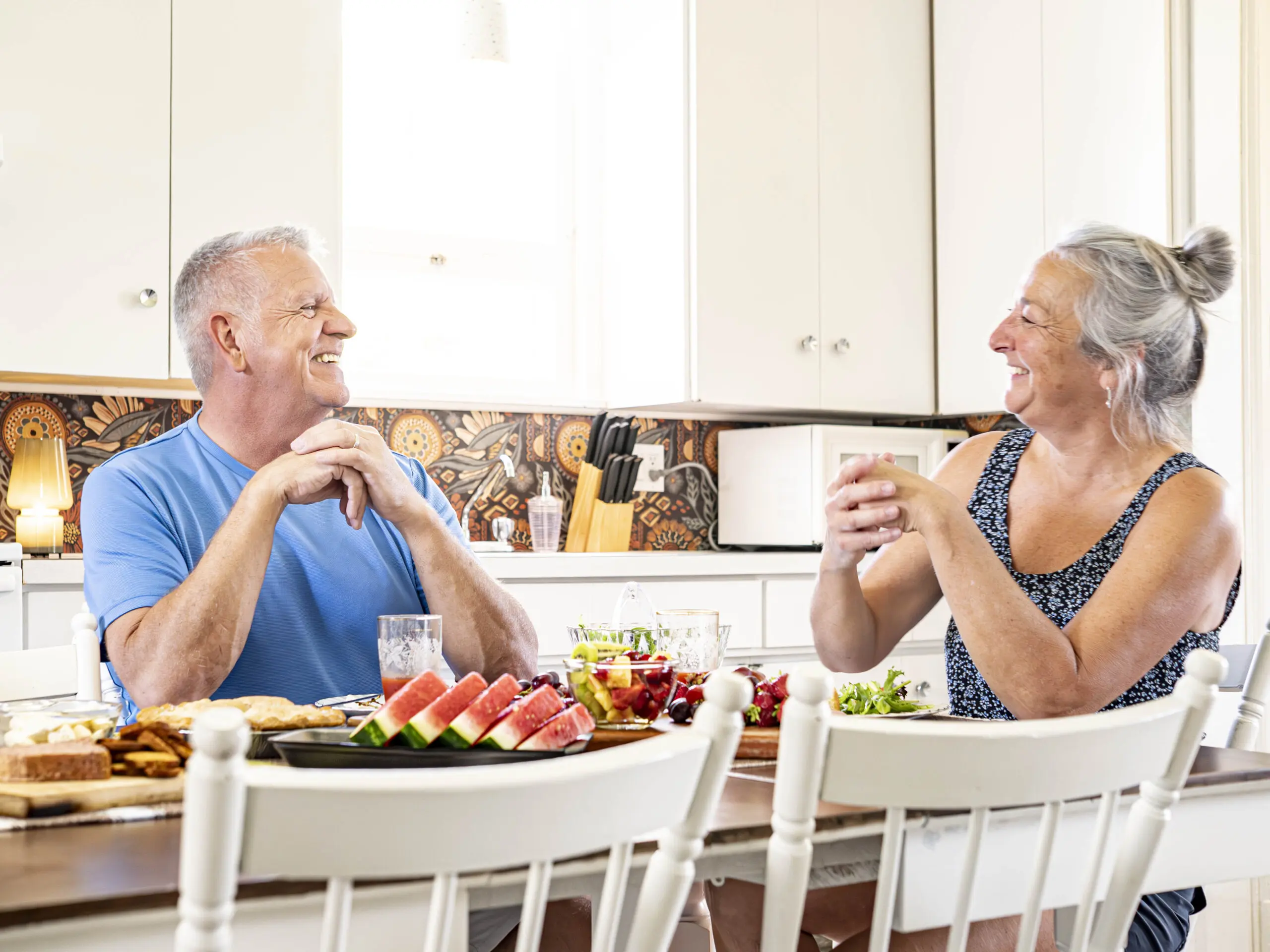 Un homme et une femme âgés sont assis à une table de cuisine, souriant et discutant. La table est garnie de divers aliments, dont de la pastèque, des fruits et du pain. La cuisine est lumineuse et moderne, avec des placards blancs.