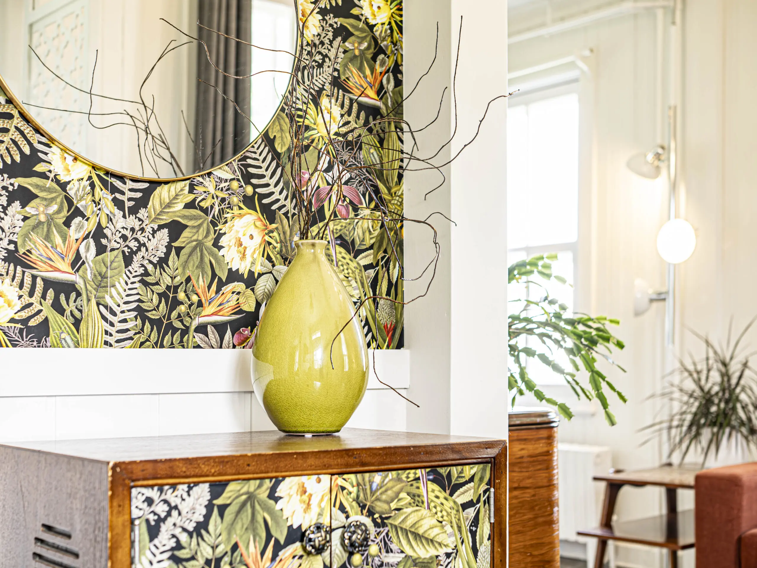 Un vase jaune-vert aux branches nues est posé sur un meuble en bois décoré d'un papier peint floral, à côté d'un miroir rond reflétant une pièce lumineuse et remplie de plantes.