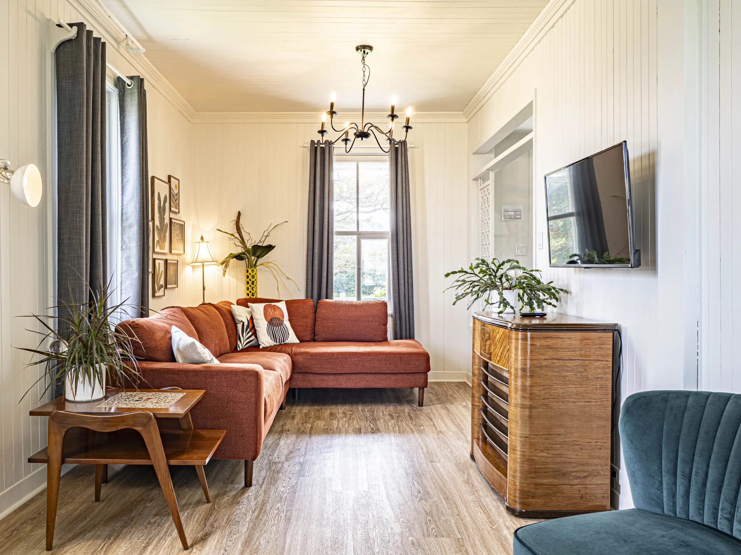 Un salon lumineux avec un canapé sectionnel orange brûlé, une table d'appoint avec des plantes, une télévision murale, un meuble en bois, une chaise verte, des œuvres d'art encadrées et une grande fenêtre avec des rideaux gris laissant entrer la lumière naturelle.