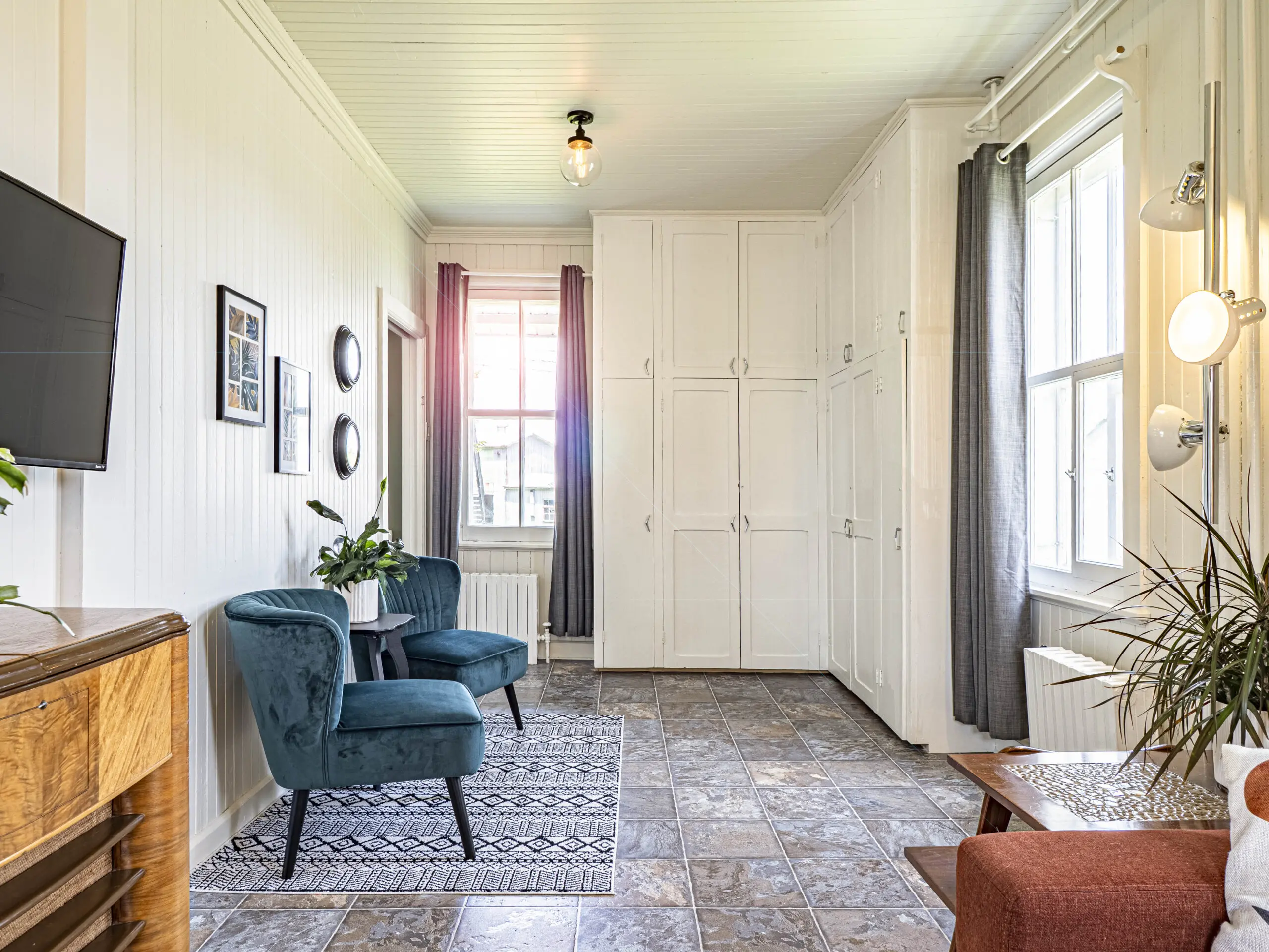 Salon lumineux avec deux fauteuils sarcelle, un tapis à motifs, une télévision murale, des armoires blanches, de grandes fenêtres avec des rideaux gris, des plantes et un lampadaire sur un sol carrelé.