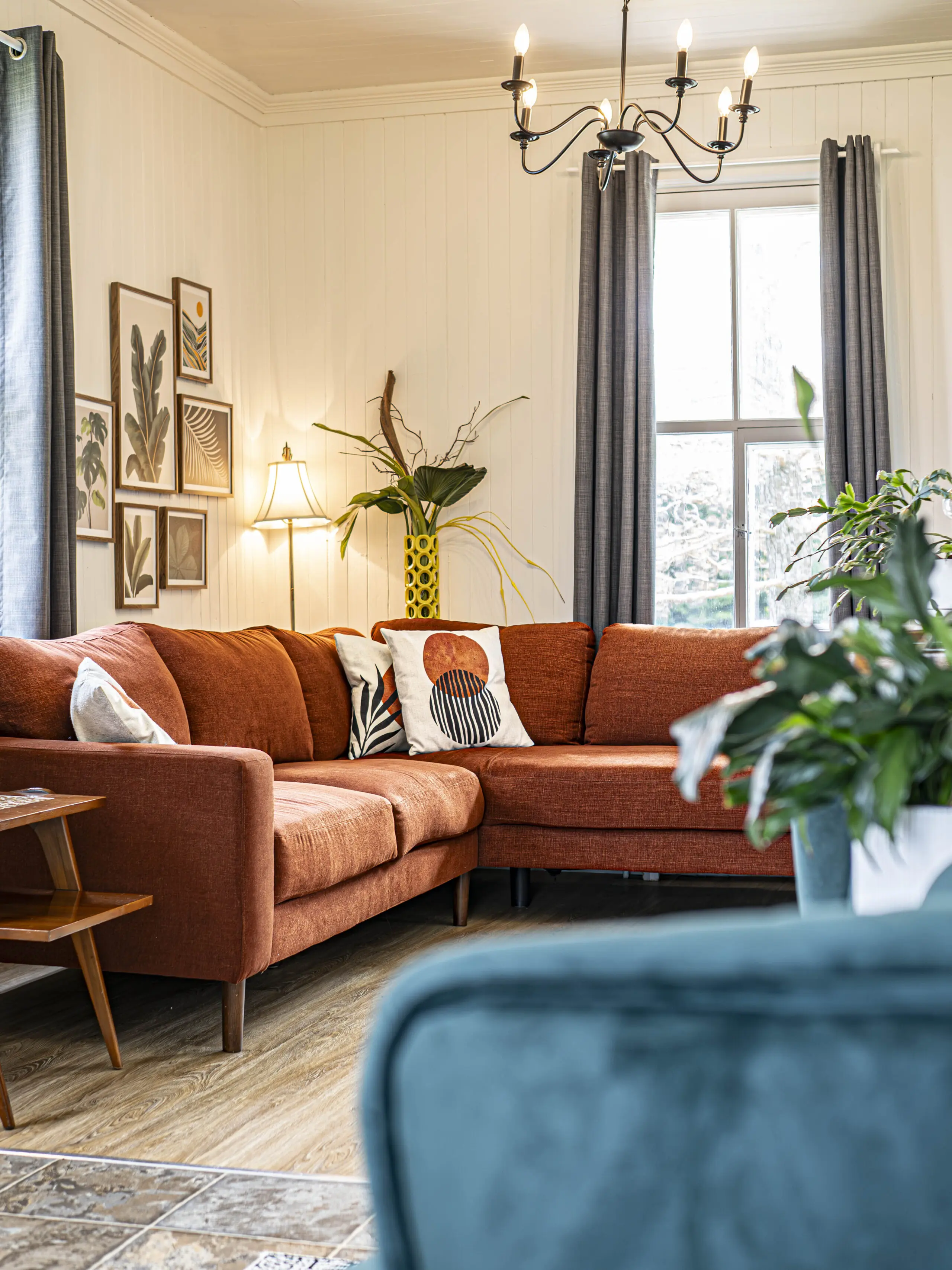 Salon confortable avec un canapé sectionnel de couleur rouille, des coussins à motifs, des œuvres d'art murales, un vase jaune avec de grandes plantes, et une grande fenêtre avec des rideaux gris. Un lustre est suspendu au plafond.