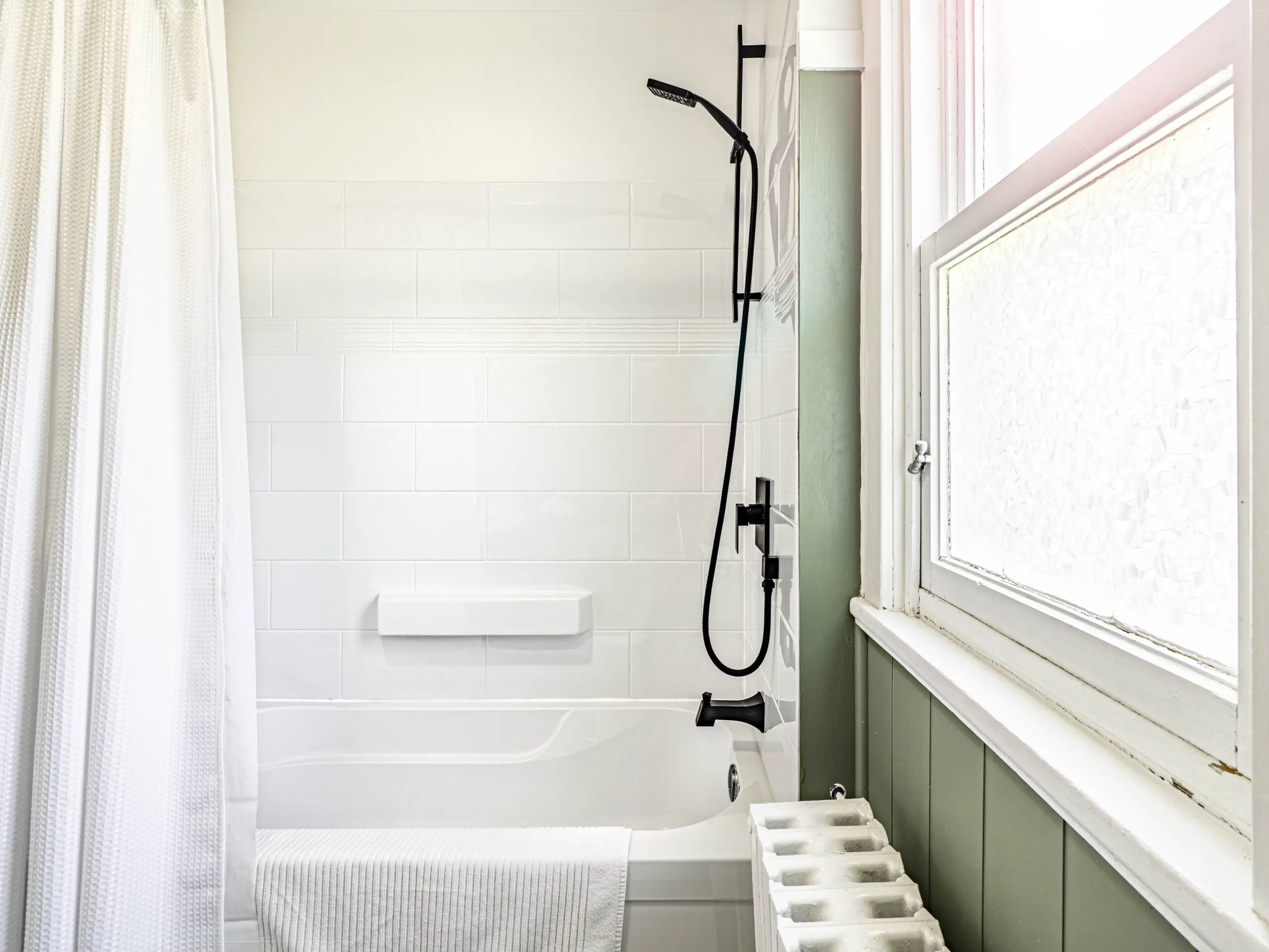 Salle de bains lumineuse avec une baignoire blanche, une douche moderne noire, des murs en carrelage blanc, un rideau de douche blanc, une grande fenêtre en verre dépoli et un radiateur sous la fenêtre. Une serviette est suspendue sur le côté de la baignoire.