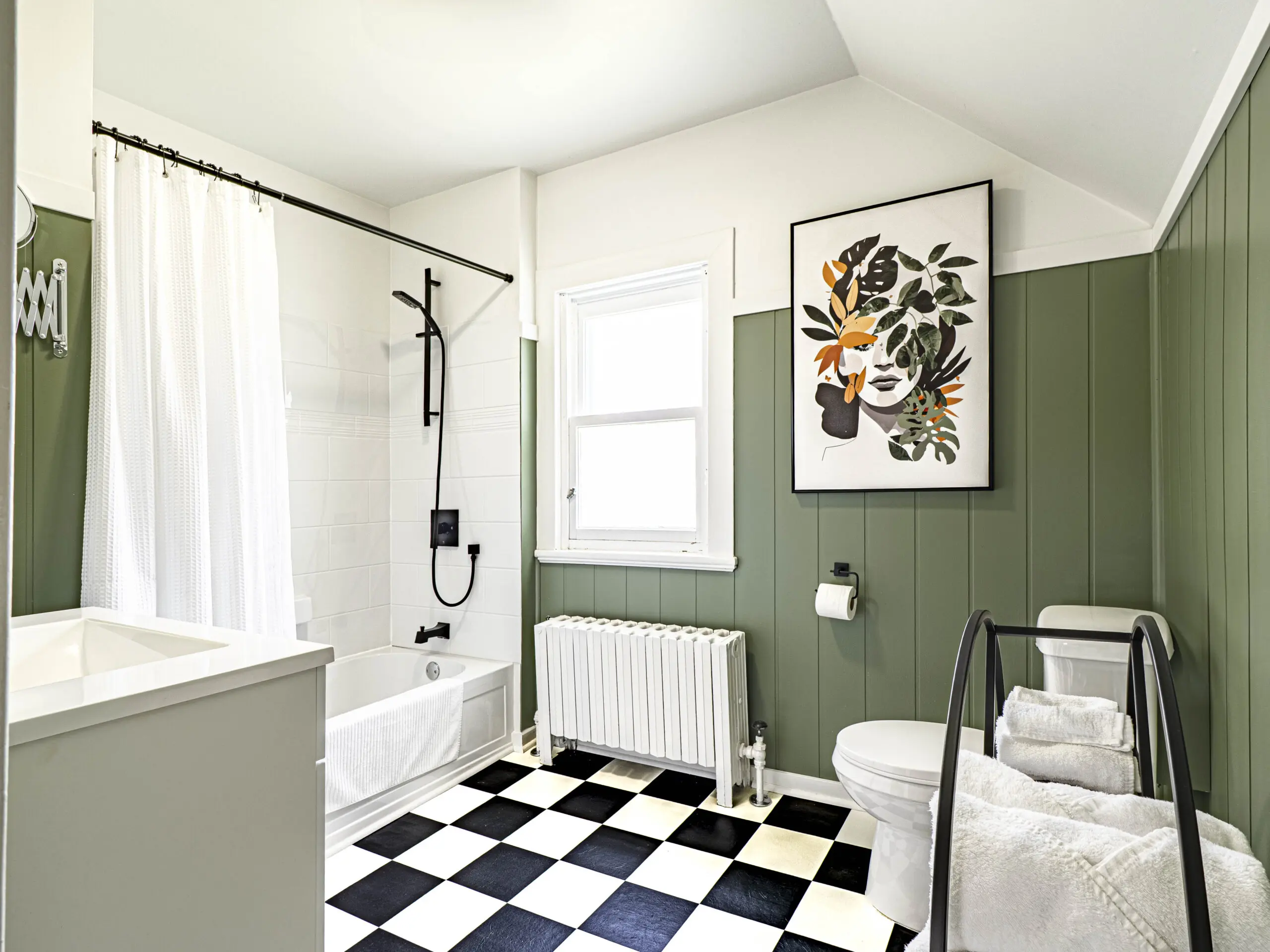 Une salle de bains moderne avec des murs lambrissés verts, une baignoire blanche avec une douche noire, une fenêtre, un sol à carreaux noirs et blancs, des toilettes, un porte-serviettes et une œuvre d'art murale représentant un visage avec des feuilles colorées.