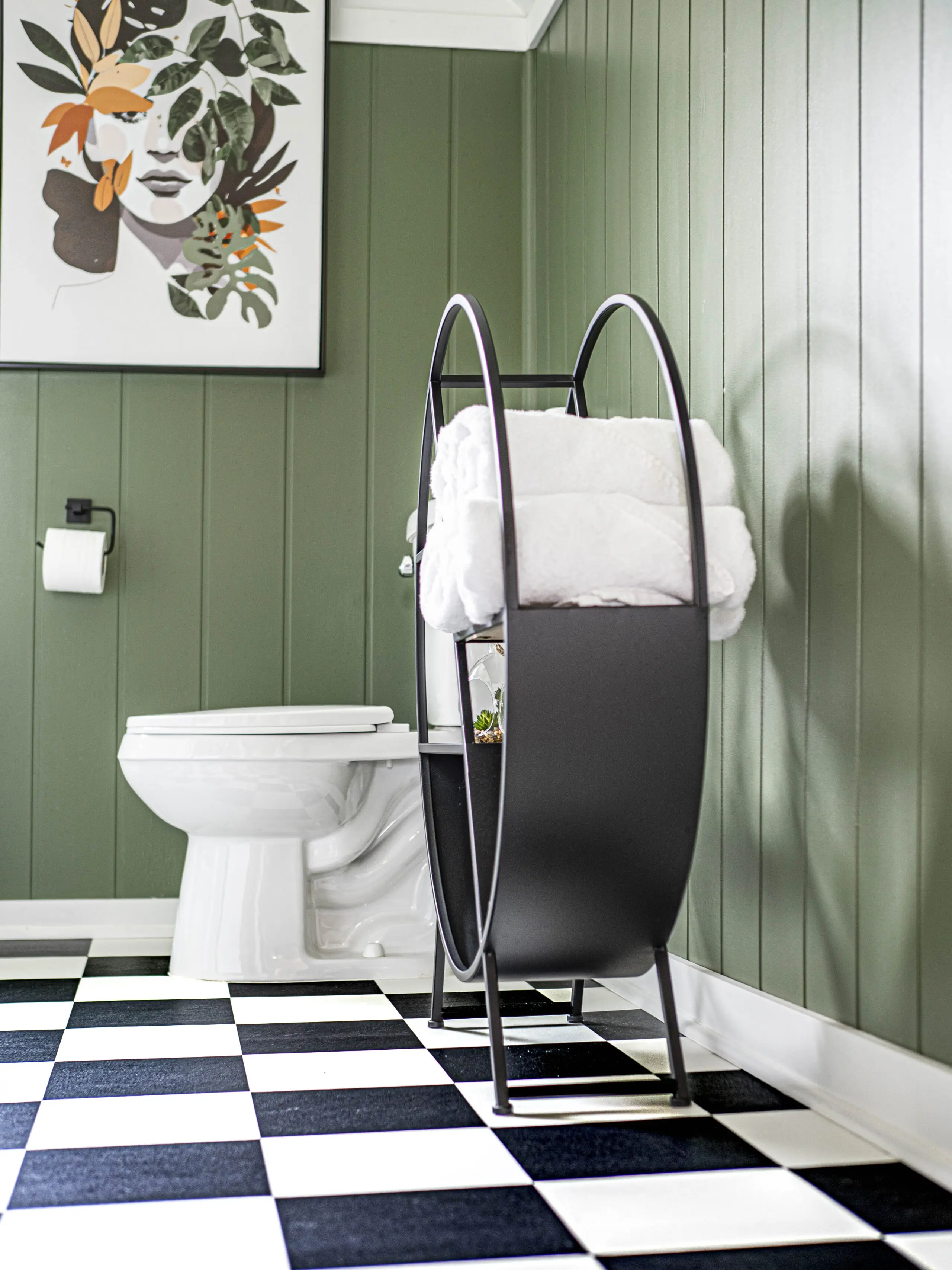 Une salle de bains aux murs lambrissés de vert, avec des toilettes blanches, un porte-papier noir, un sol à carreaux noirs et blancs, un porte-serviettes noir contenant des serviettes blanches et un portrait d'art moderne encadré sur le mur.
